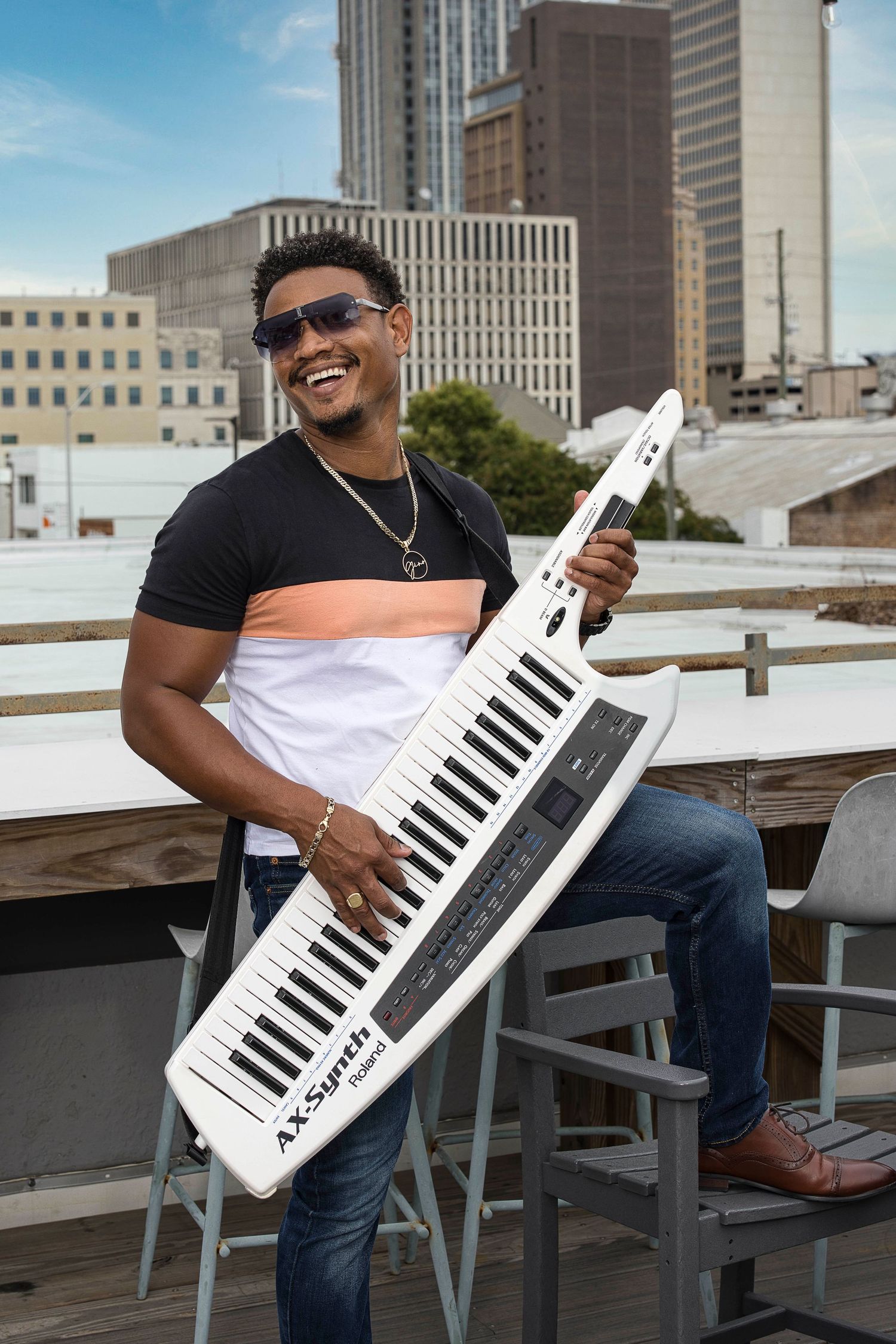 A musician poses with a white keytar instrument on an urban rooftop setting.