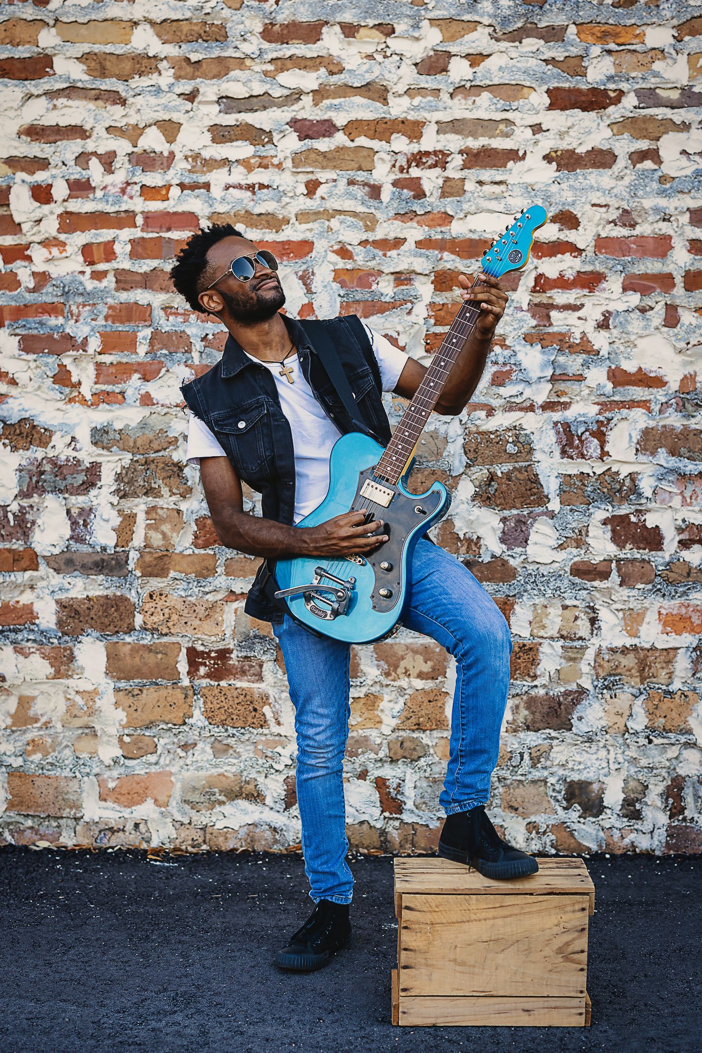 A musician performs with a blue electric guitar while standing on a wooden box against brick wall.