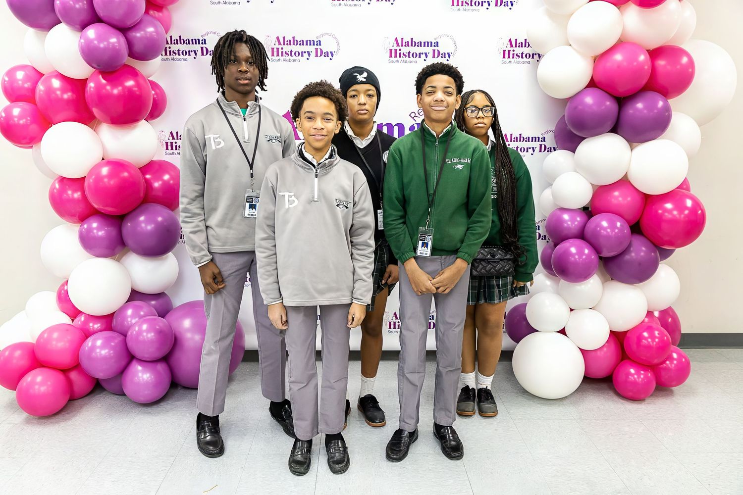 Group of students in uniforms posing in front of balloon arch with school event backdrop.