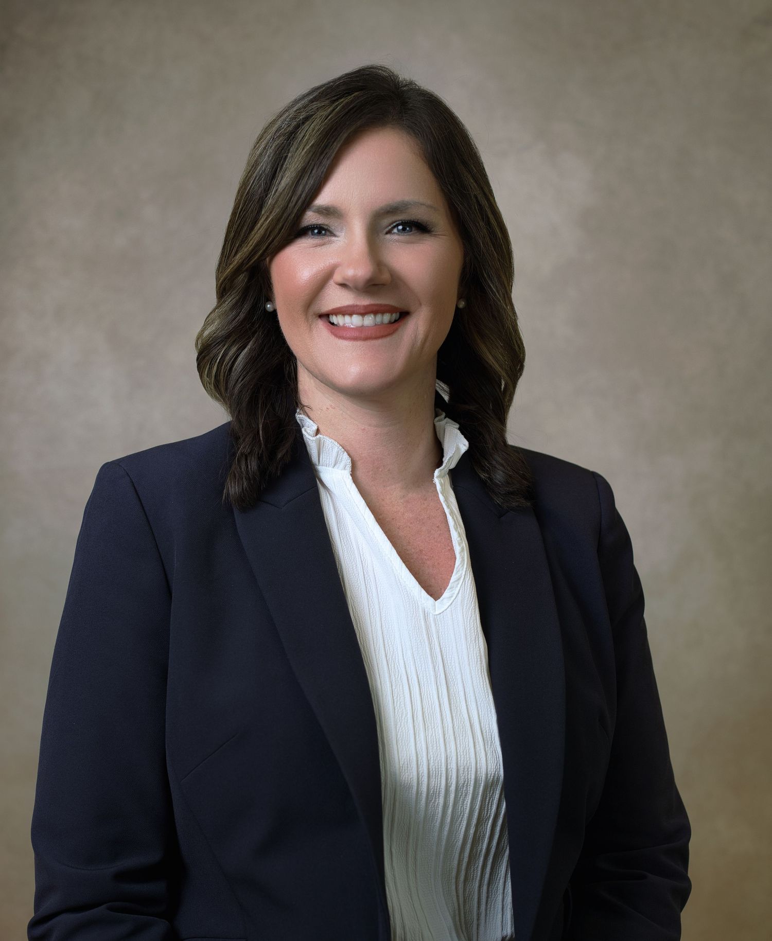 Professional headshot of woman in navy blazer and white blouse against beige background.