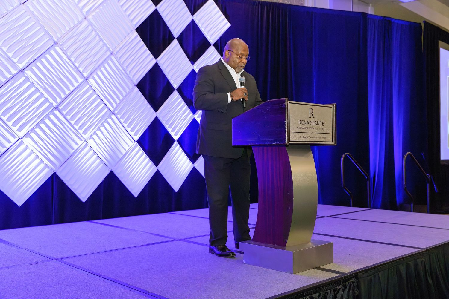 A man in a suit speaks at a podium on a decorated stage with blue and white diamond patterned backdrop.