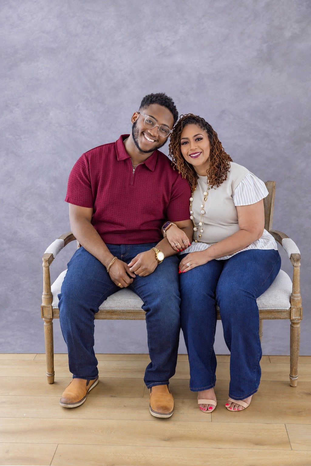 A smiling couple sits close together on a wooden bench against a gray backdrop, he in a maroon polo and she in a white top and jeans.