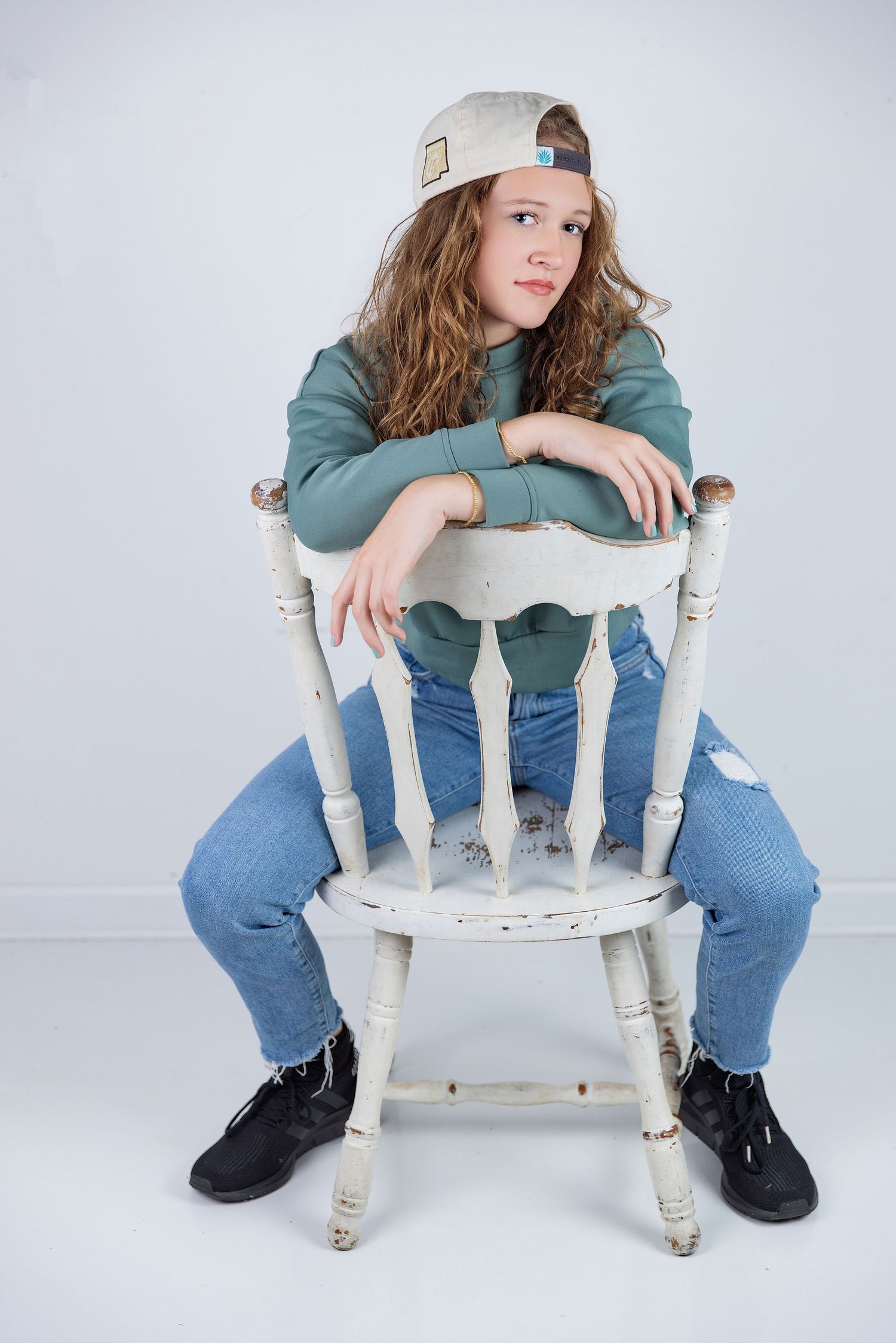 Young girl in green sweatshirt, ripped jeans, and backward cap sits on a rustic white wooden chair in a studio.