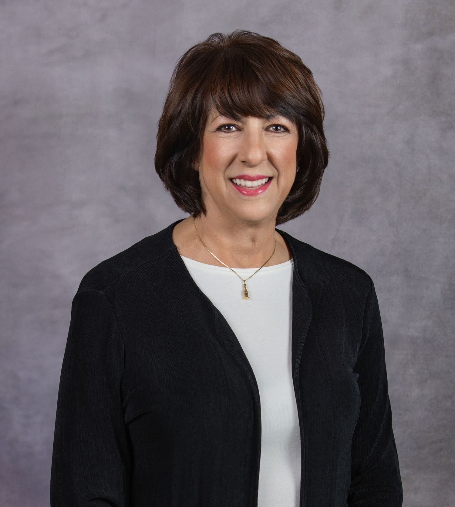 Professional headshot portrait of someone in a black blazer and white top with a warm smile against a gray backdrop.