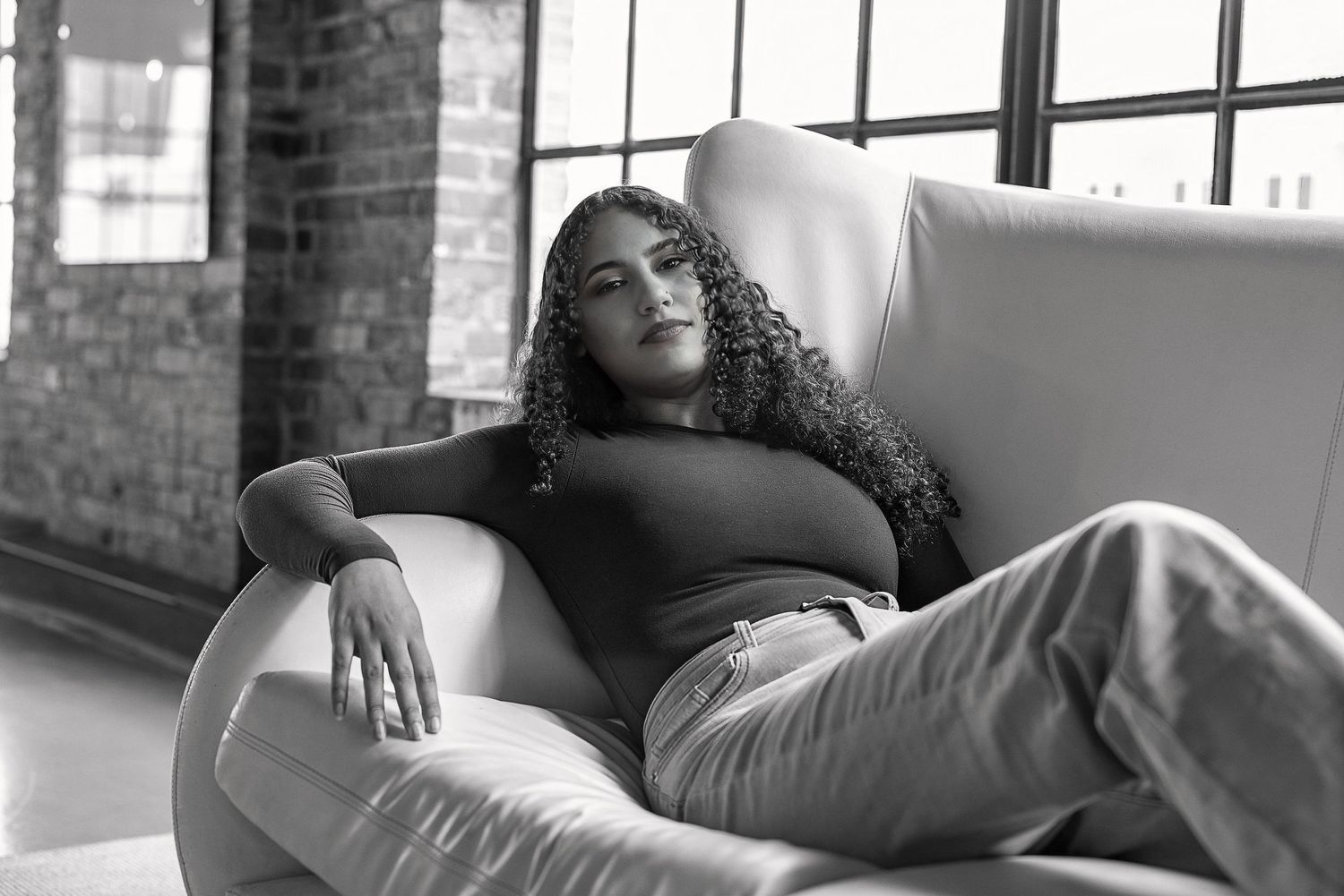 Black and white portrait of a person relaxing on a white couch near industrial windows.
