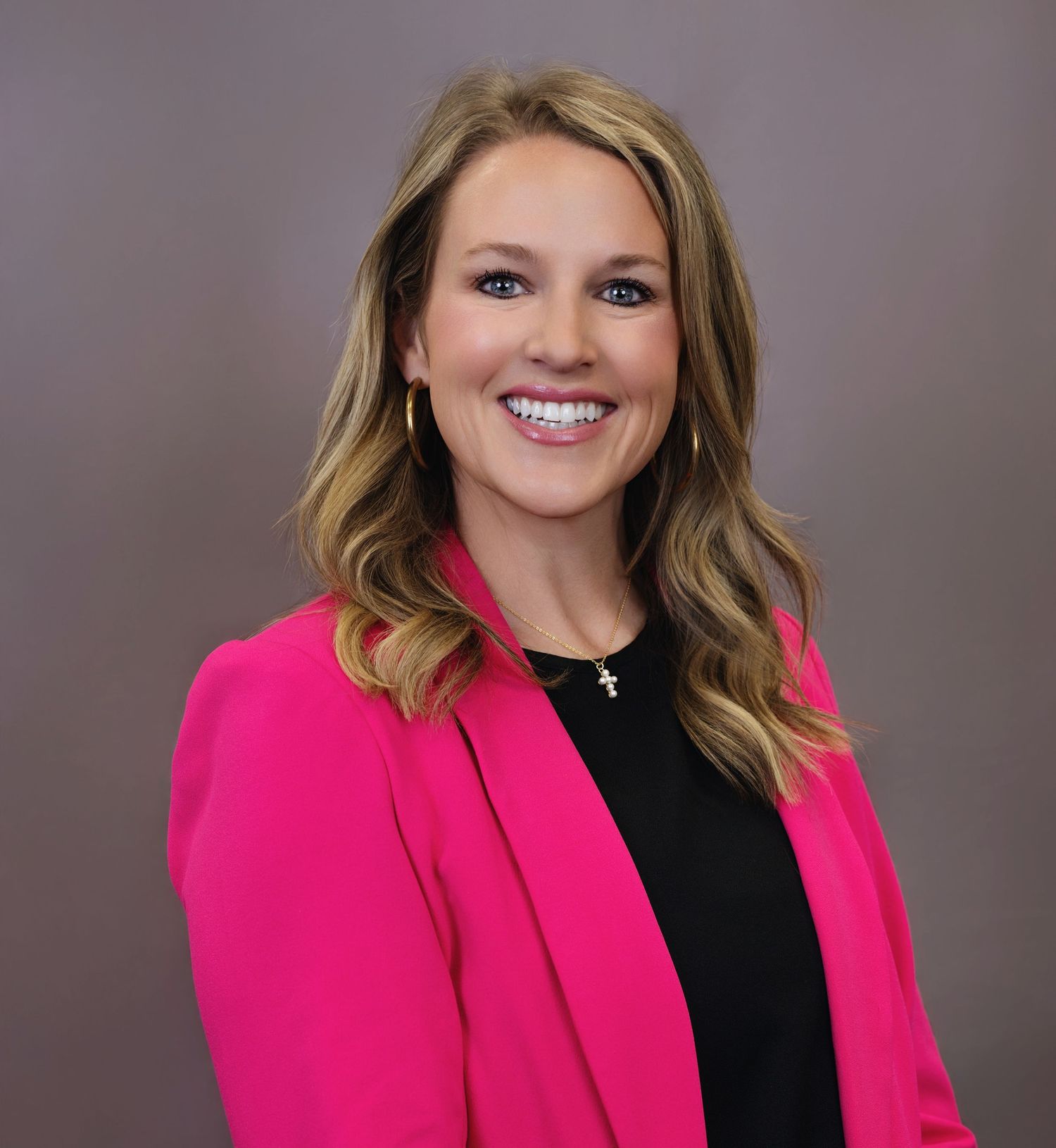 Professional headshot of someone wearing a bright pink blazer and black top with styled wavy hair smiling at the camera.