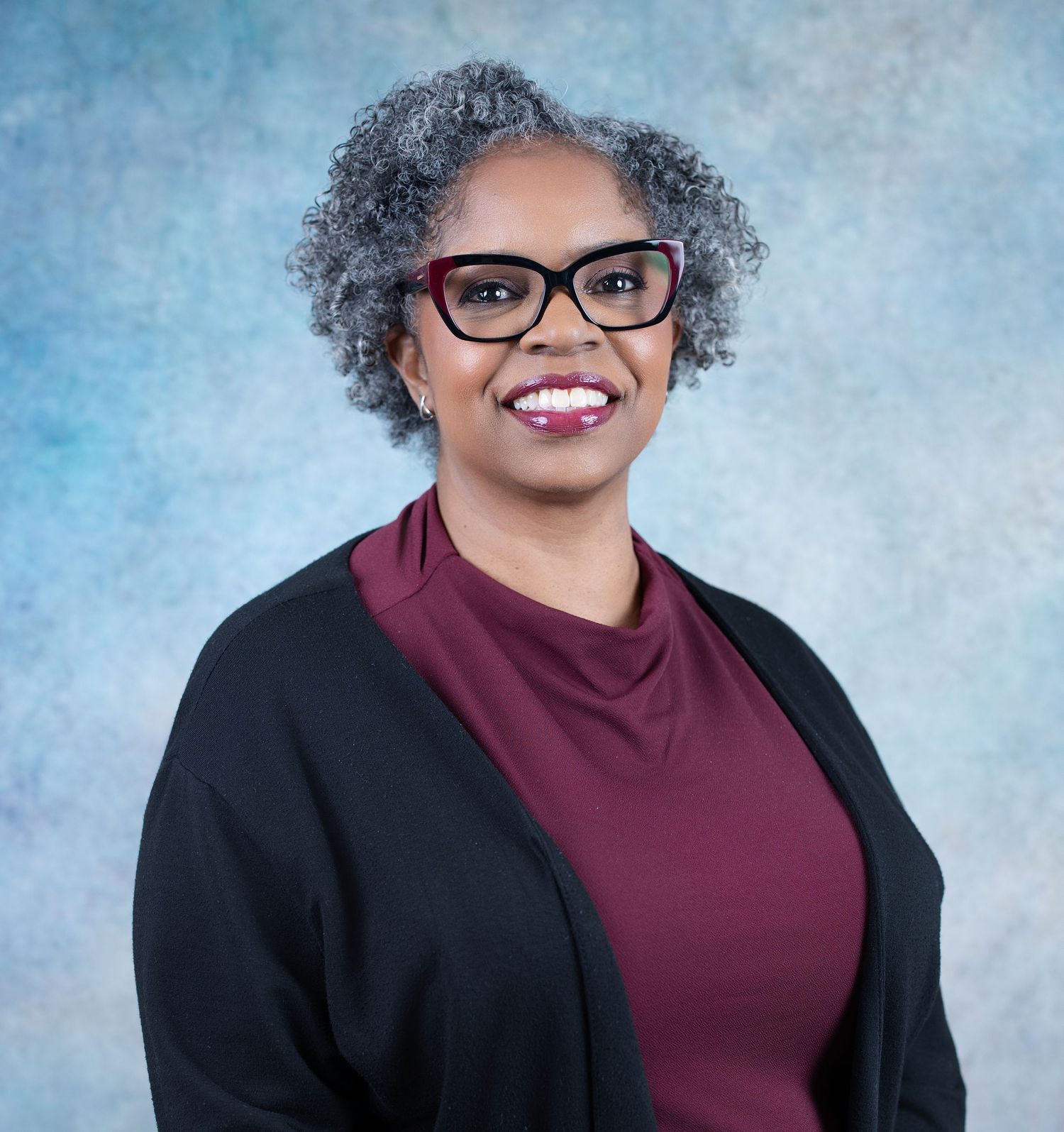 Professional headshot of a smiling woman with gray curly hair and red glasses wearing a burgundy top and black cardigan.