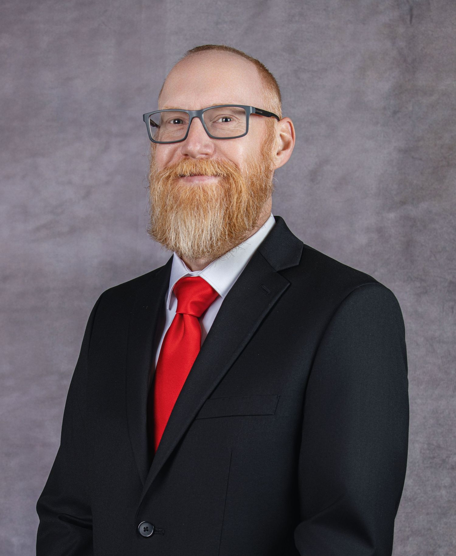 Professional headshot of person wearing black suit jacket, red tie and glasses with a ginger beard against grey background.