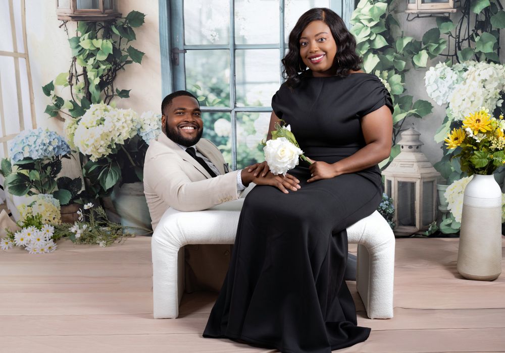 Elegant couple in black dress and beige suit pose together on white seating with floral decor and greenery.