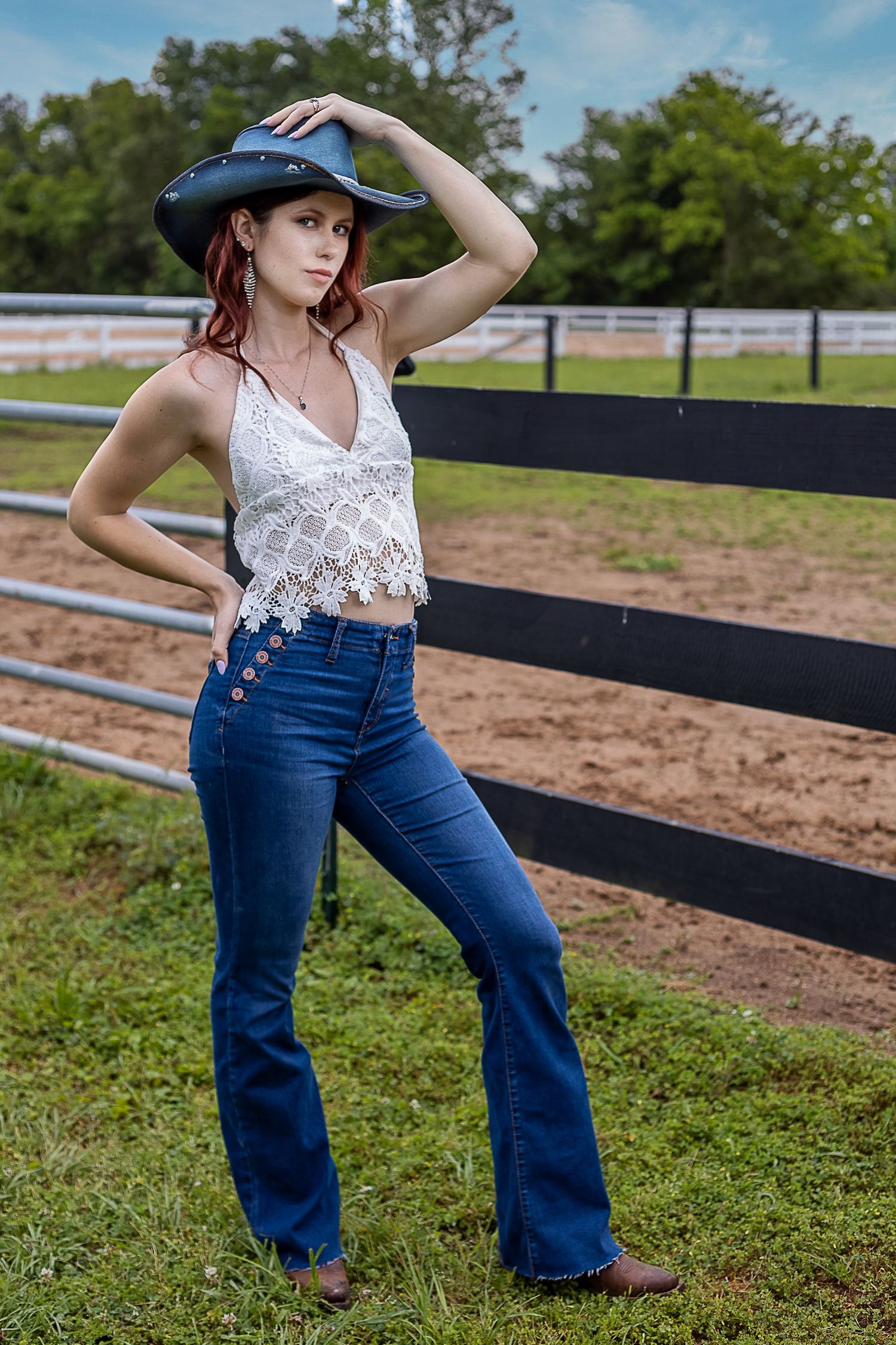 A woman in a lace top and flared jeans poses near a horse fence wearing an equestrian helmet.