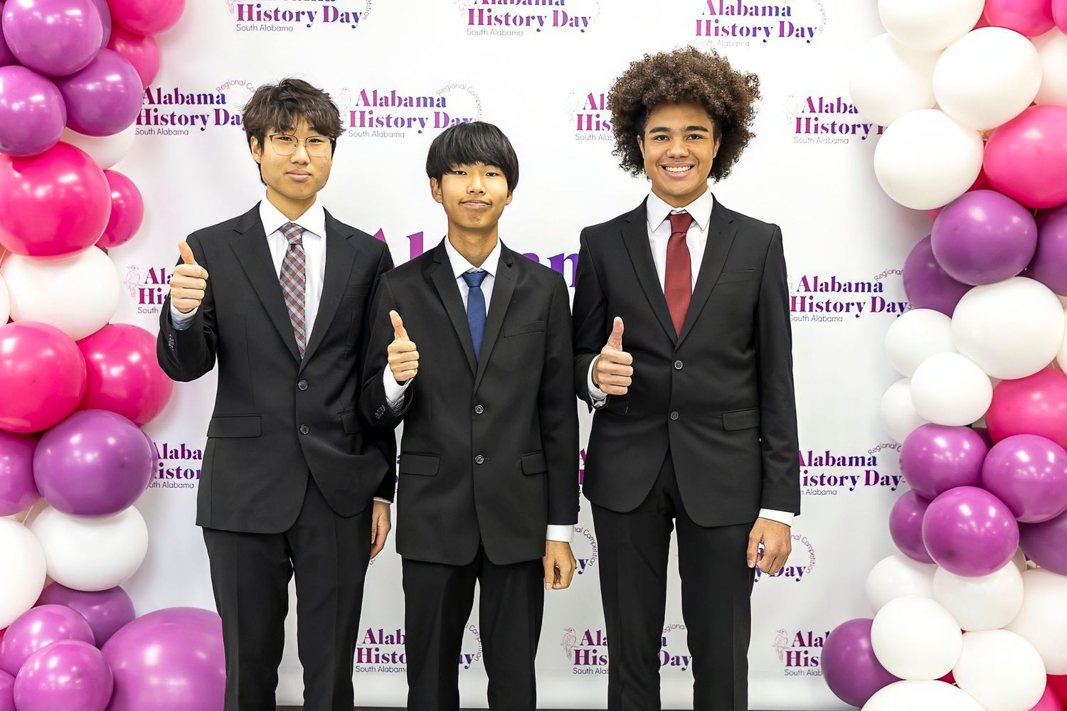 Three students in formal attire giving thumbs up in front of decorated balloon backdrop.
