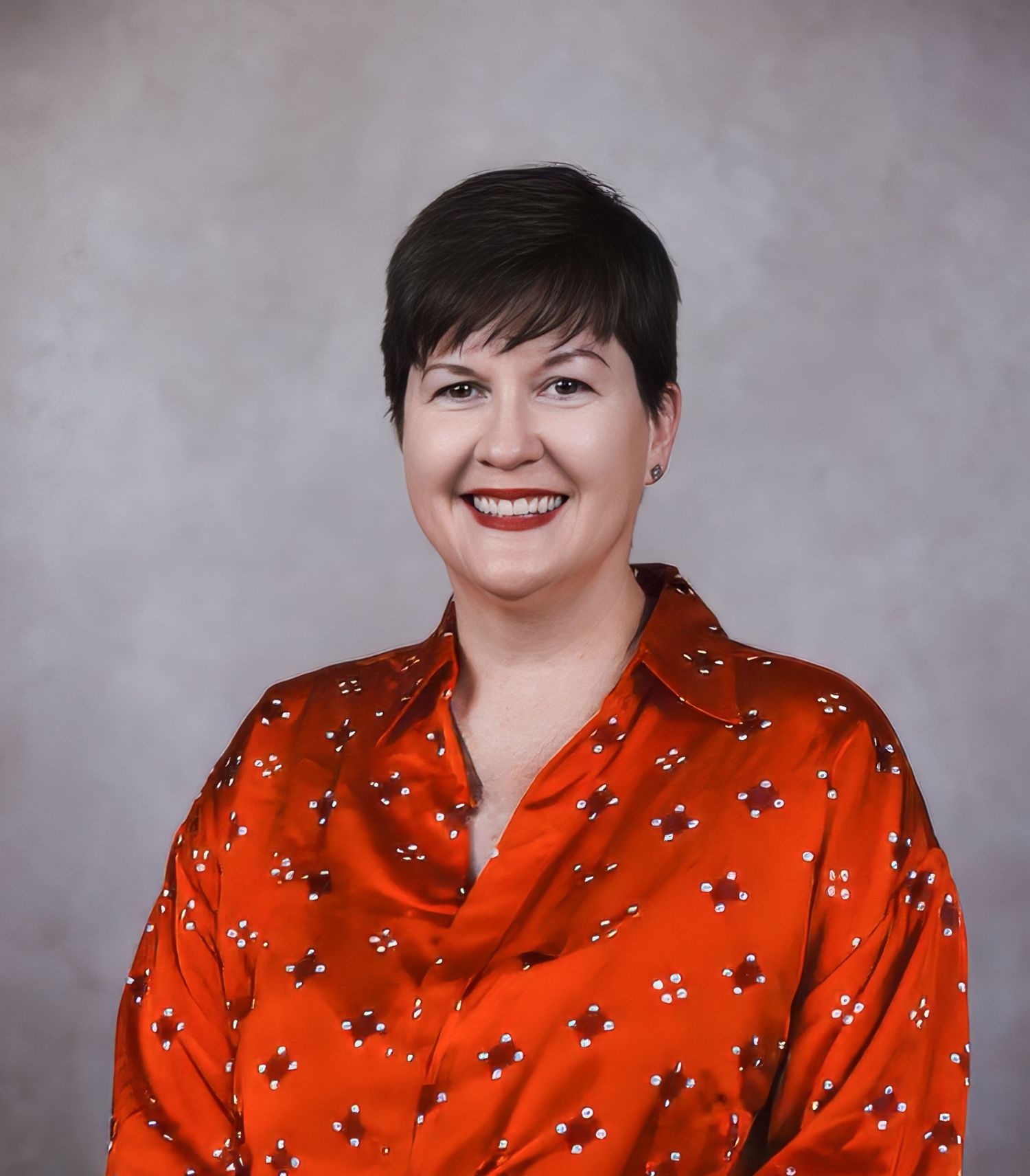 Professional studio headshot of a smiling person wearing a bright red patterned blouse against a gray background.