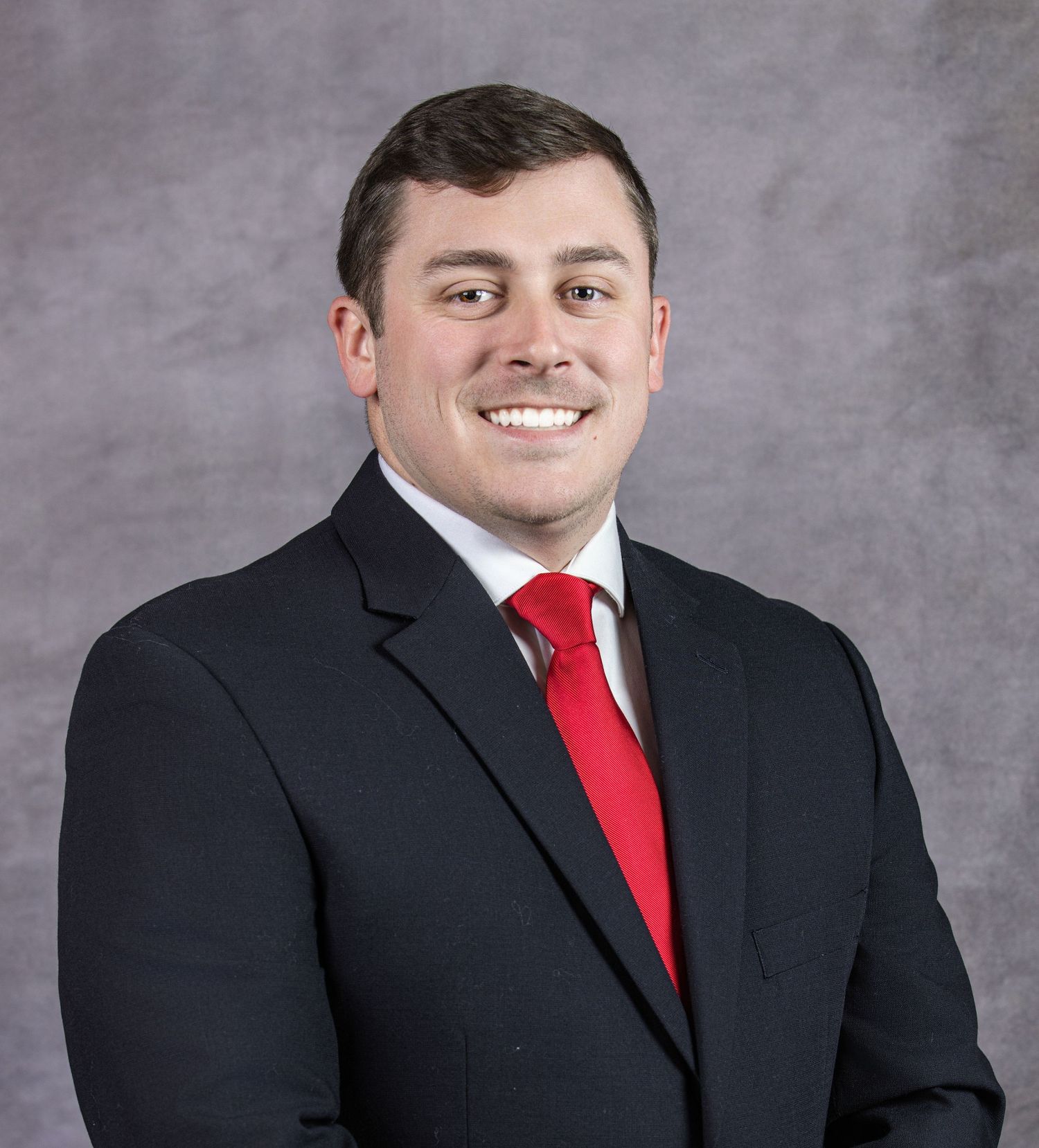 Professional headshot of someone in a black suit and red tie against a grey backdrop.