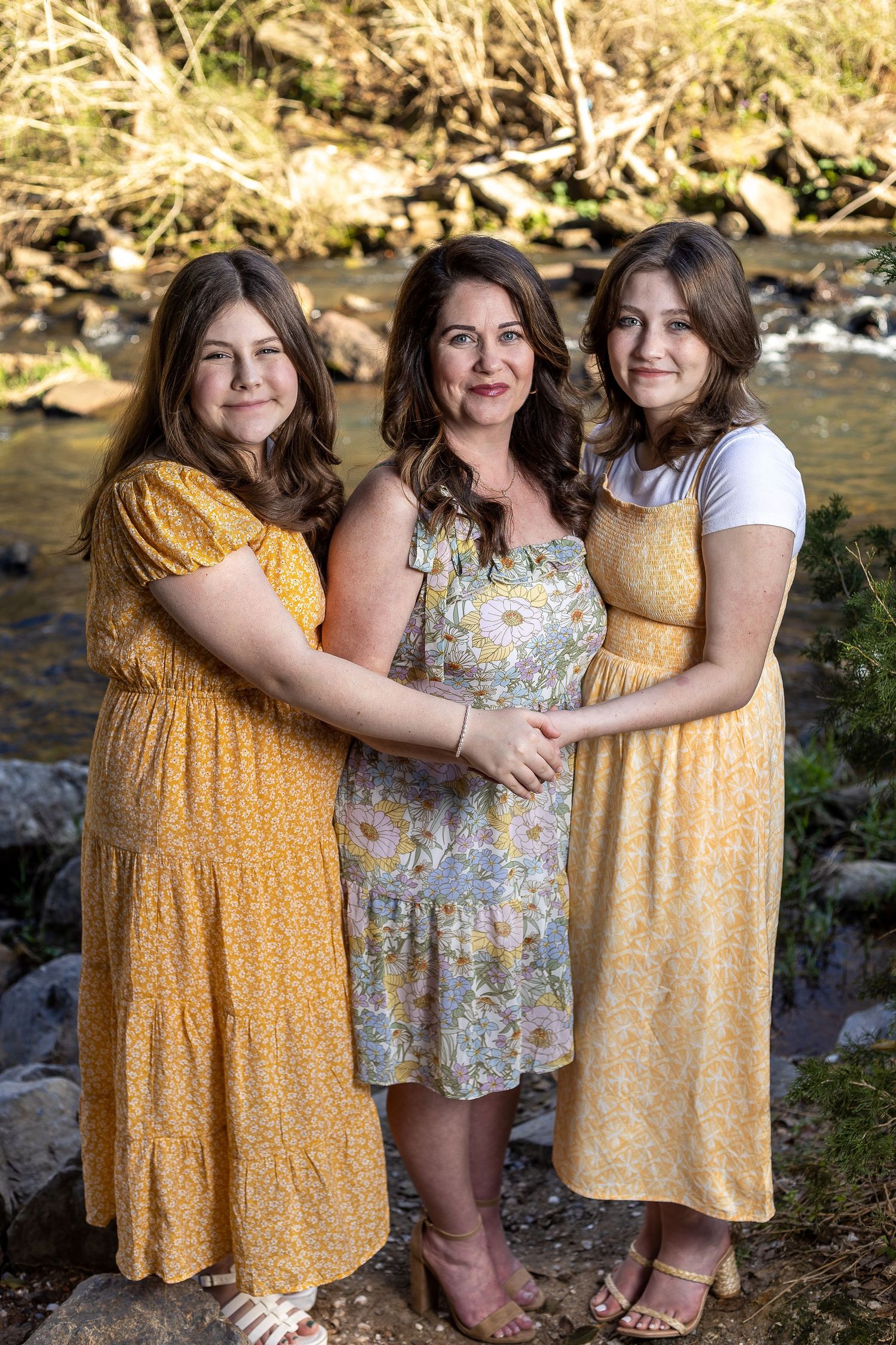 Three women in coordinating yellow and floral dresses embrace by a creek.