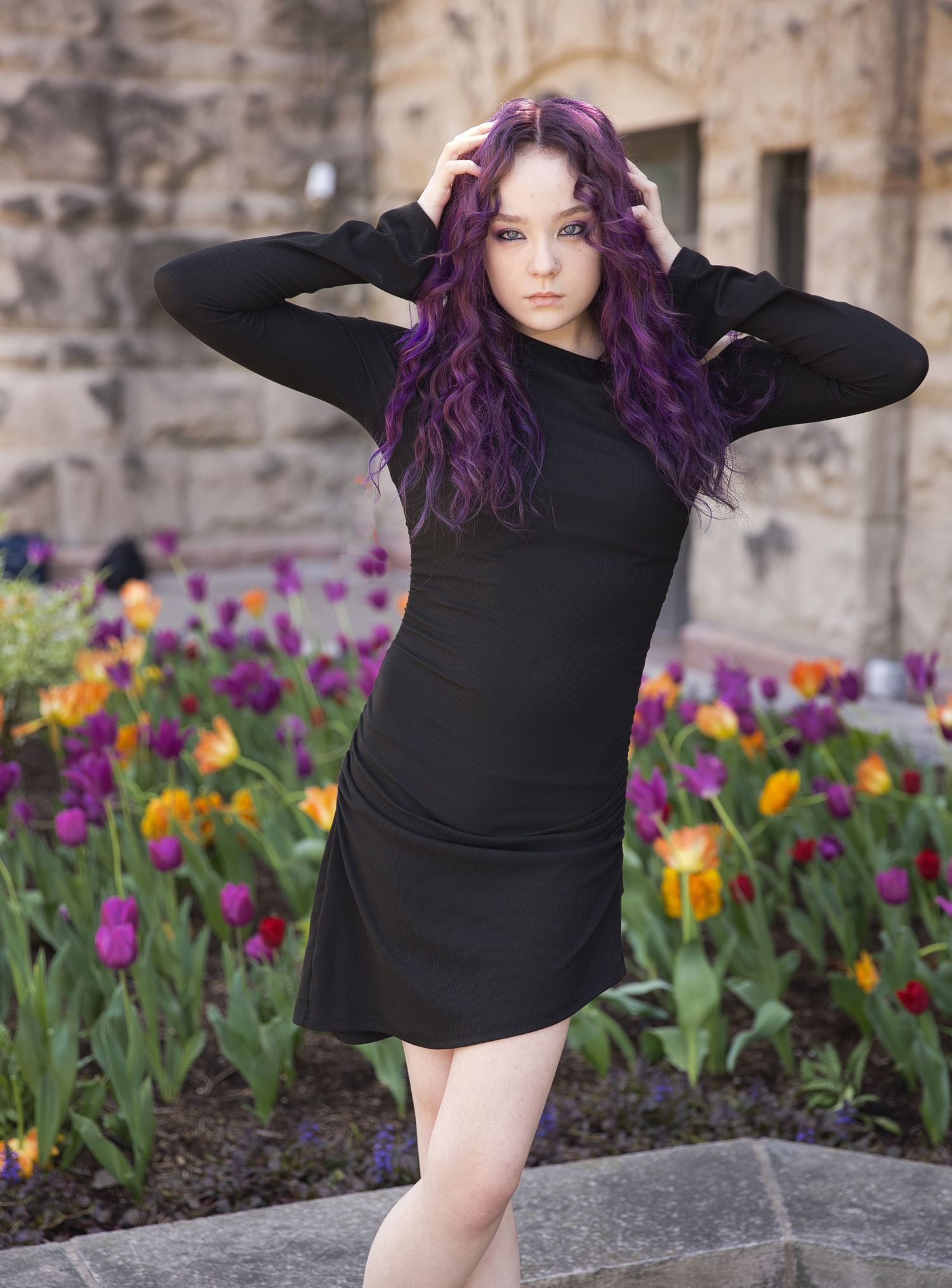 A woman in black dress poses with vibrant purple hair among colorful spring tulips.