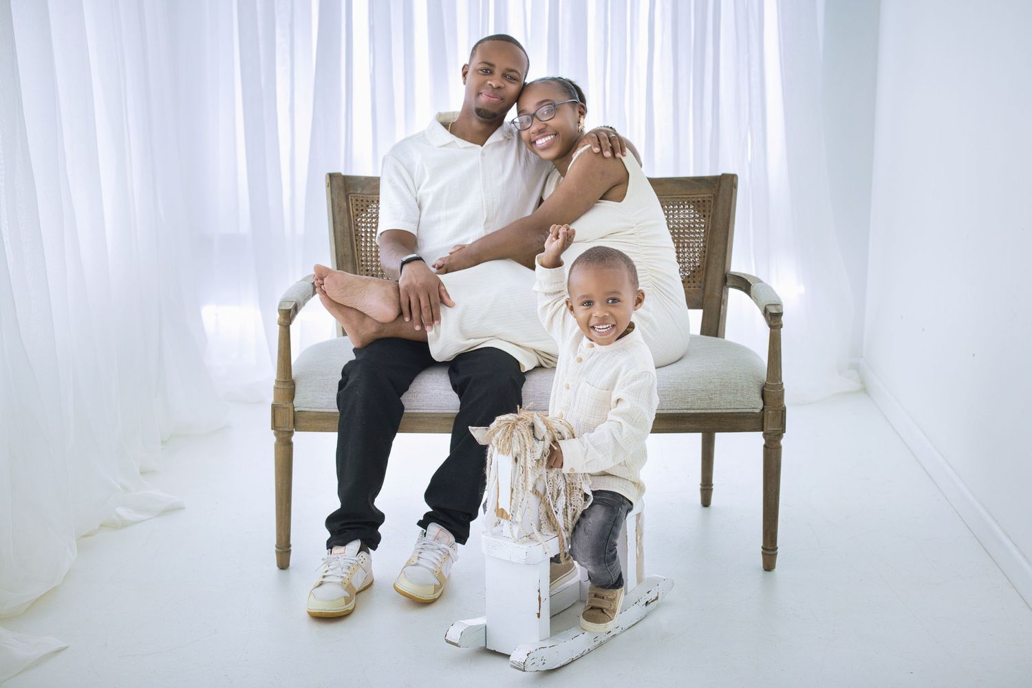 A family sits together on an elegant beige bench in a bright white studio setting against sheer curtains.
