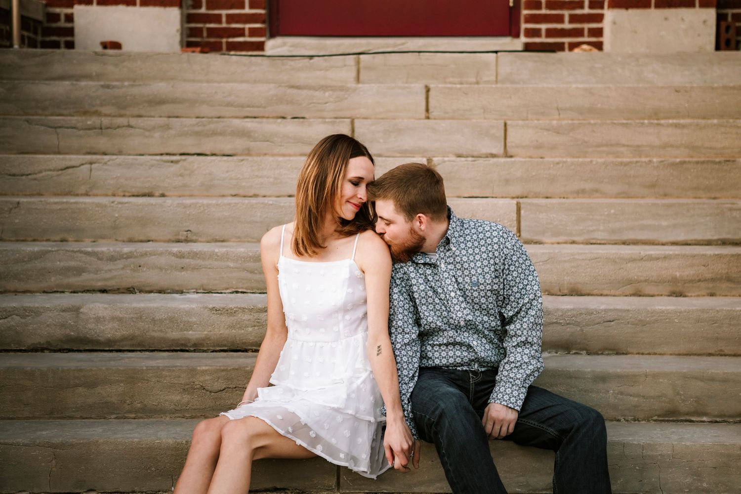 A couple sits close together sharing an intimate moment on concrete steps outside a brick building.