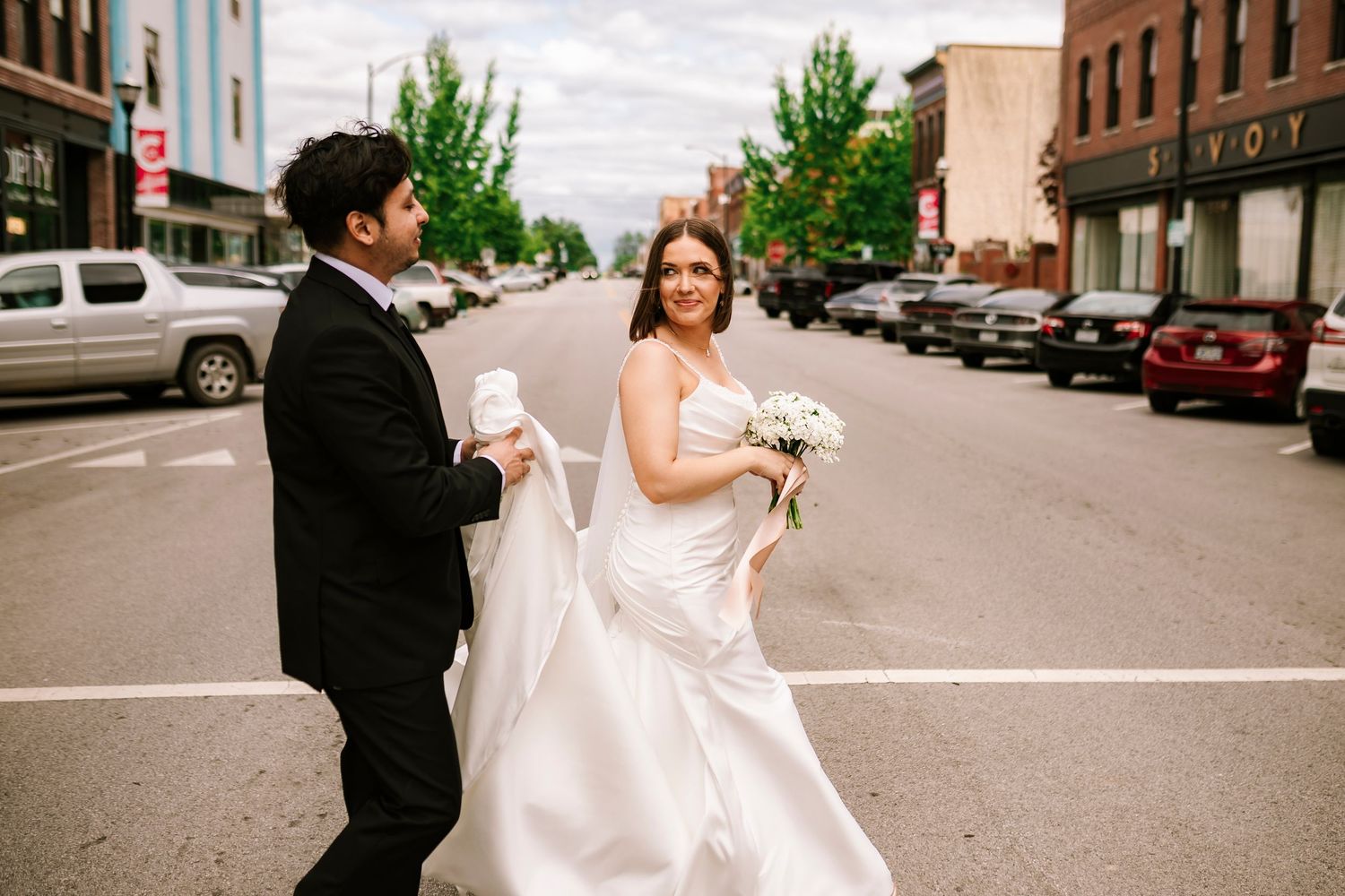 A couple in wedding attire share a candid moment walking down a street  and parked cars headed to Savoy Ballroom downtown Springfield MO.