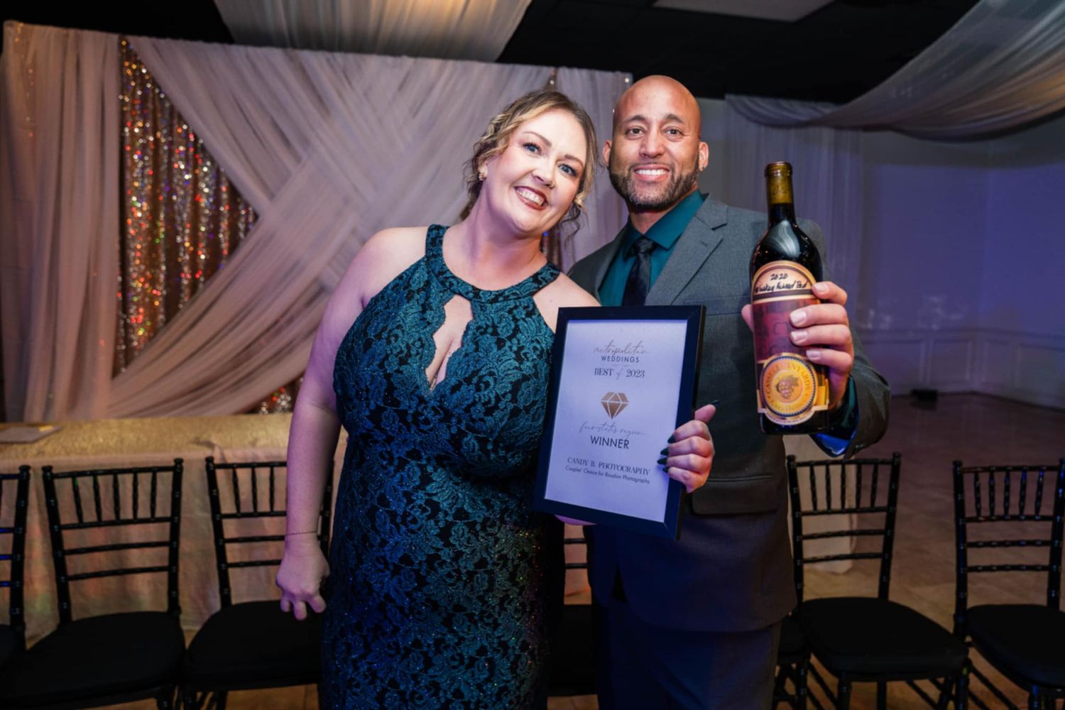 Two people pose together at an event while holding an award certificate and bottle of champagne.
