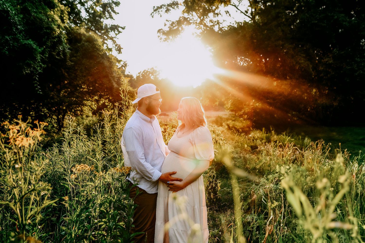 A couple shares a tender moment at sunset in a wildflower meadow with golden sunlight streaming through trees.
