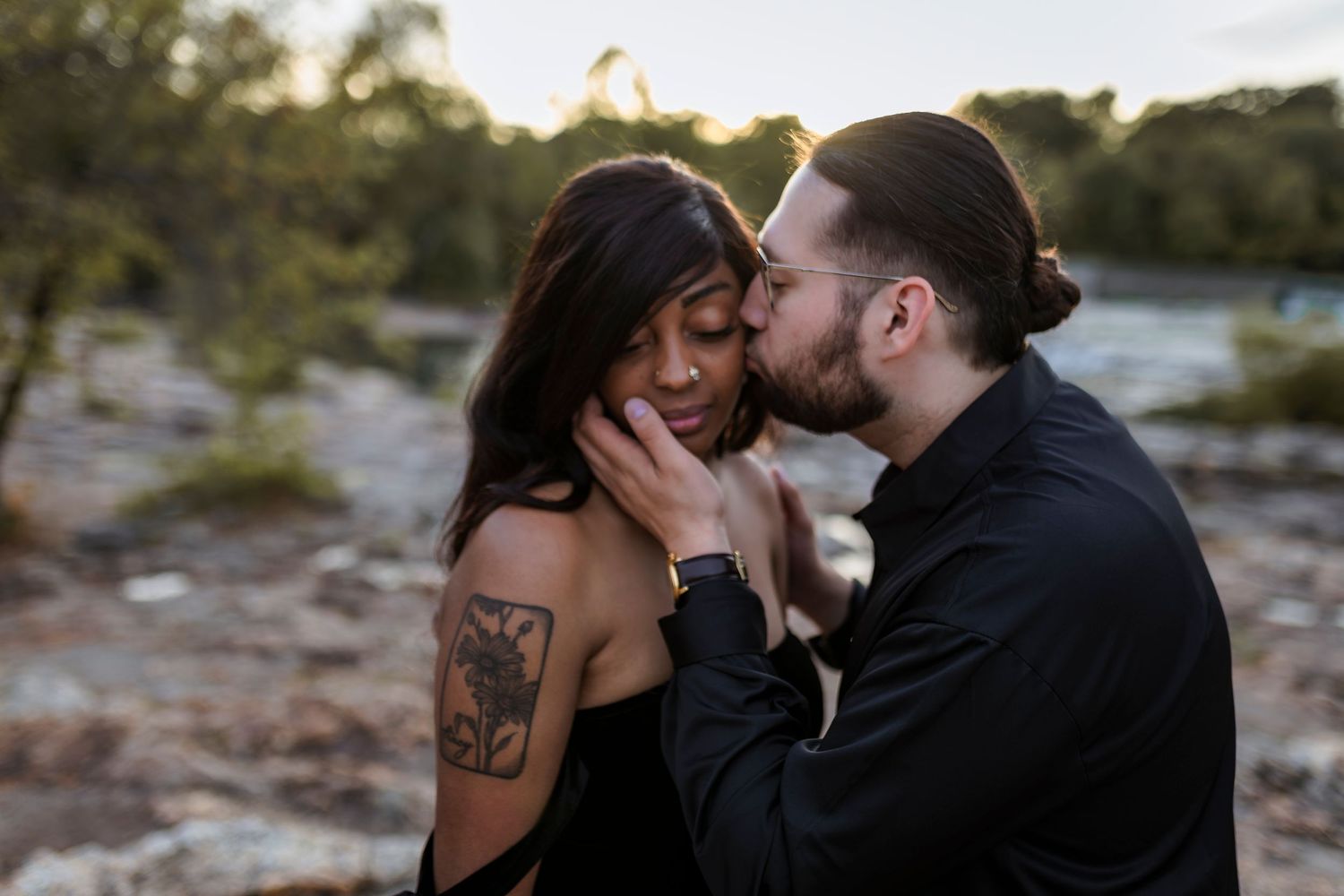 A couple share an intimate romantic moment outdoors at dusk, with one person displaying a visible tattoo on their arm.