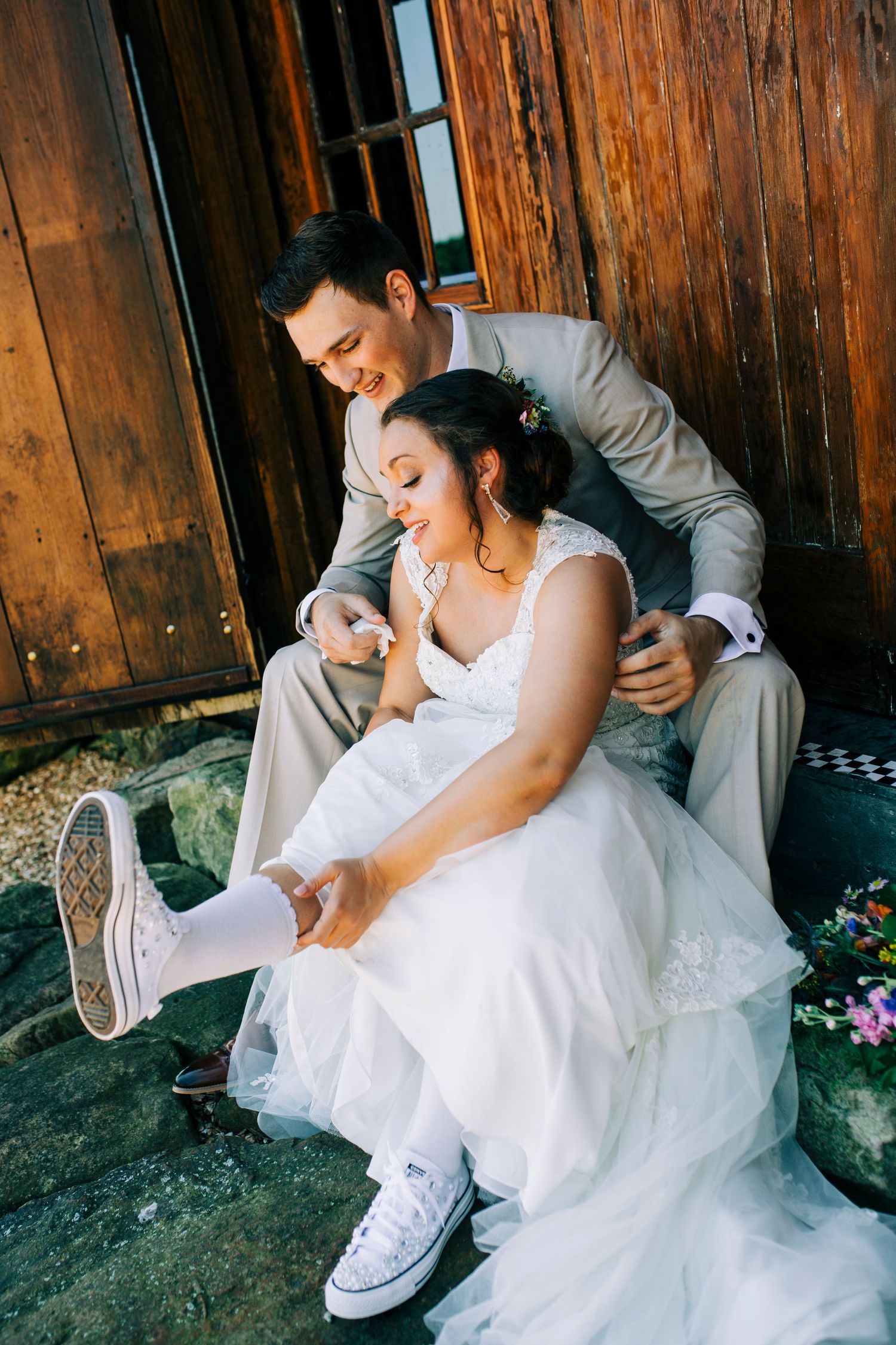 Bride in white dress and sneakers sitting with groom against rustic wooden wall at Storybook Barn in Missouri.