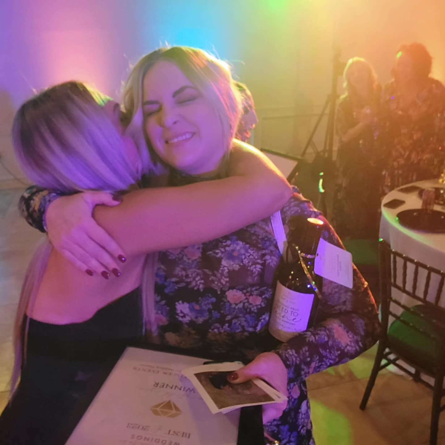 Two people share an emotional hug at an event with colorful lighting and a rainbow lens flare effect in the background.