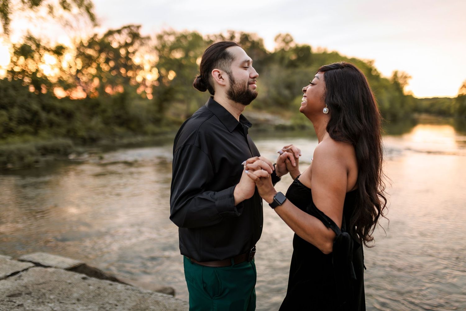 A couple shares a romantic moment at sunset by a river as they dance together in elegant attire.