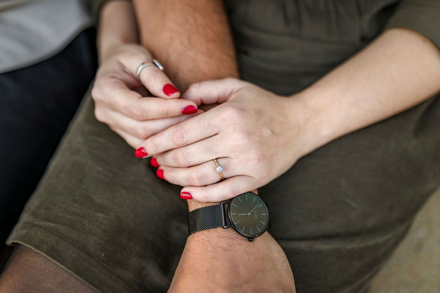 A person wearing a black watch rests their hands in their lap, displaying bright red nail polish and rings.