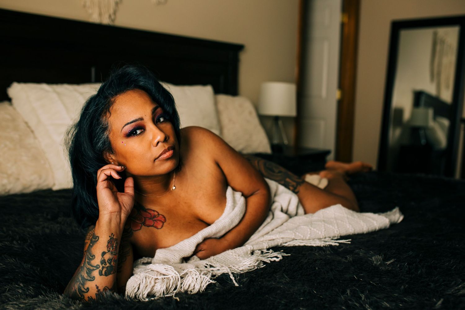 A person poses seductively on dark bedding while wearing white lingerie and showcasing artistic tattoos in moody lighting.