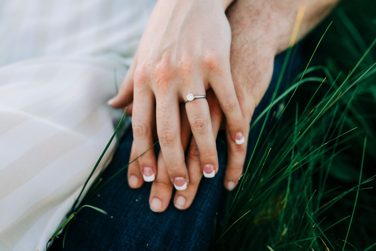 Two hands with wedding rings tenderly touch on dark fabric with green plant leaves in soft natural lighting.