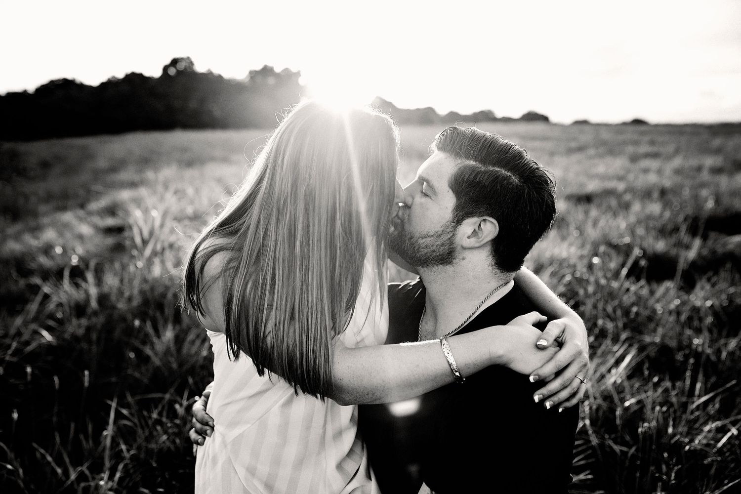 Black and white photo of a romantic couple embracing in a sunlit field at golden hour.
