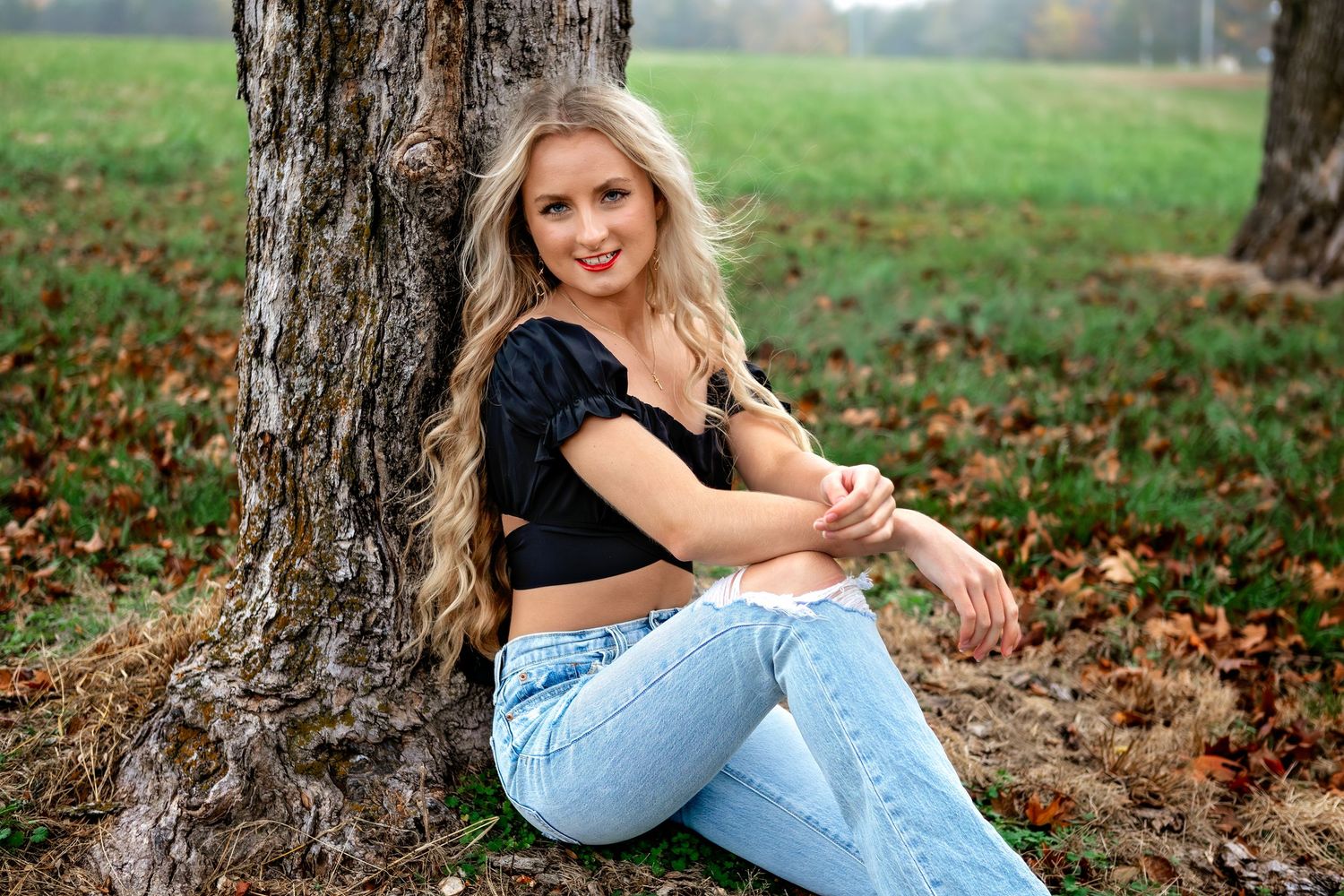 Casual outdoor fall portrait shows woman in black top and jeans sitting against tree trunk with autumn leaves on ground.