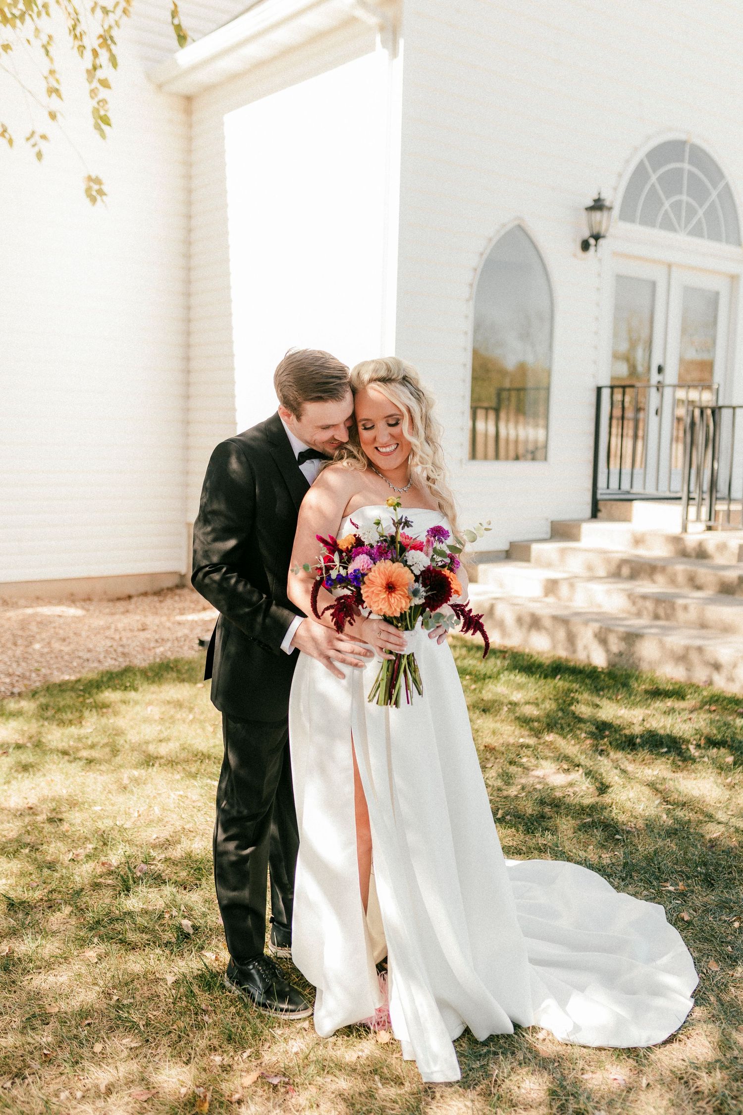 Romantic newlyweds share an intimate moment outdoors as groom embraces bride holding vibrant floral bouquet in front of white chapel.