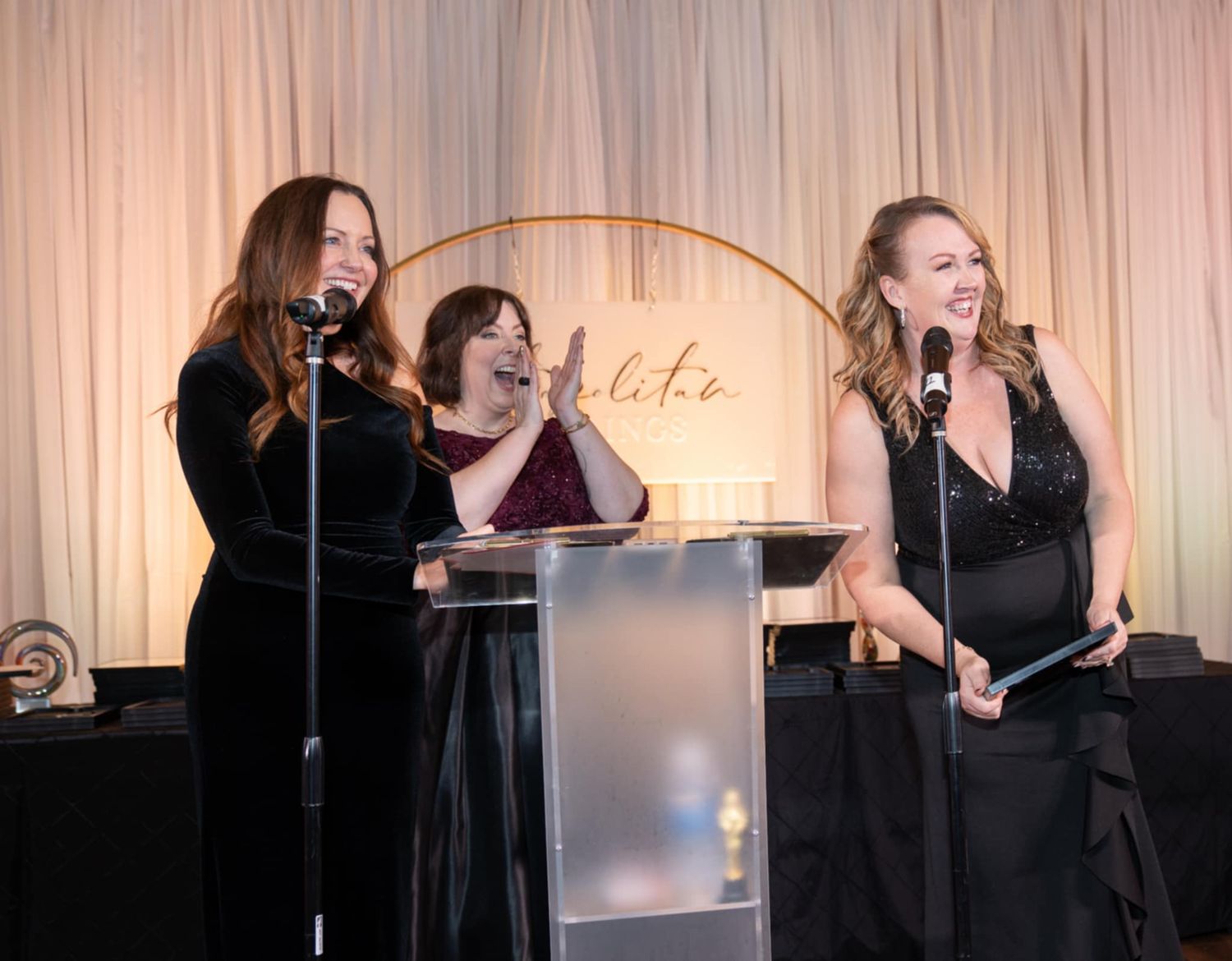 A group wearing matching black outfits performs together against a beige curtain backdrop in a professional setting.