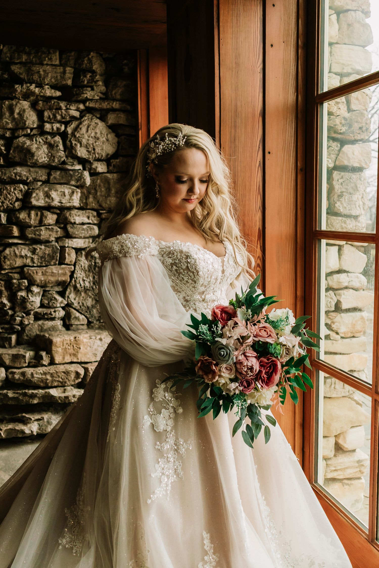Bride in flowing white lace gown with sheer sleeves holds romantic pink and burgundy bouquet near rustic stone wall and window.