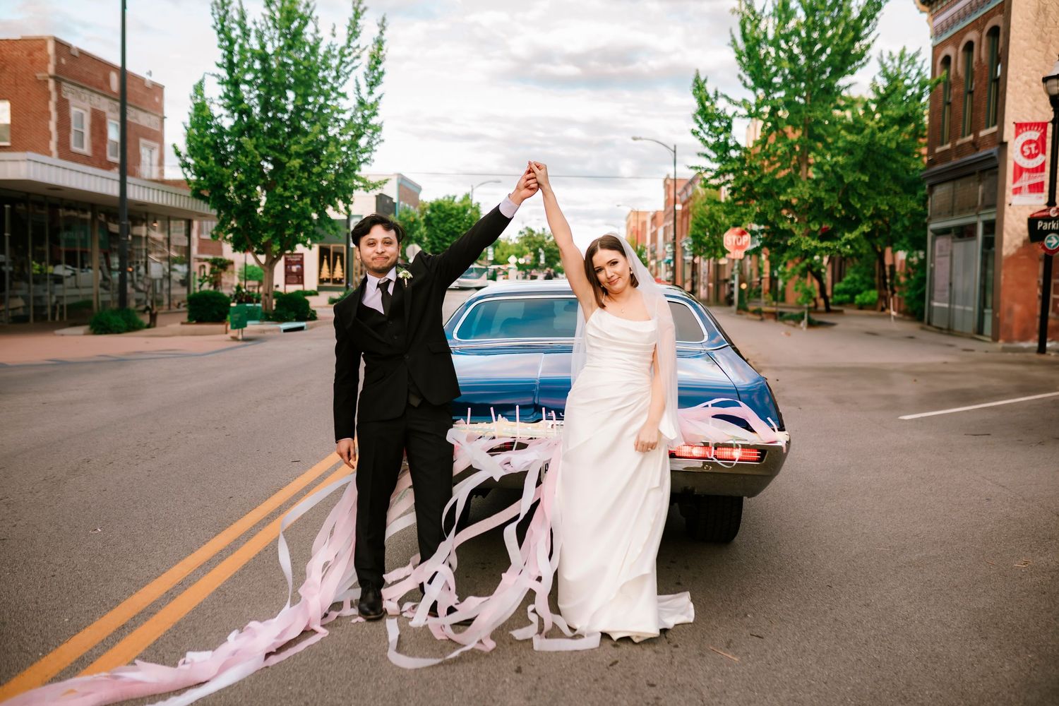 A couple standing by a vintage blue car on a downtown street with decorated trailing ribbons.
