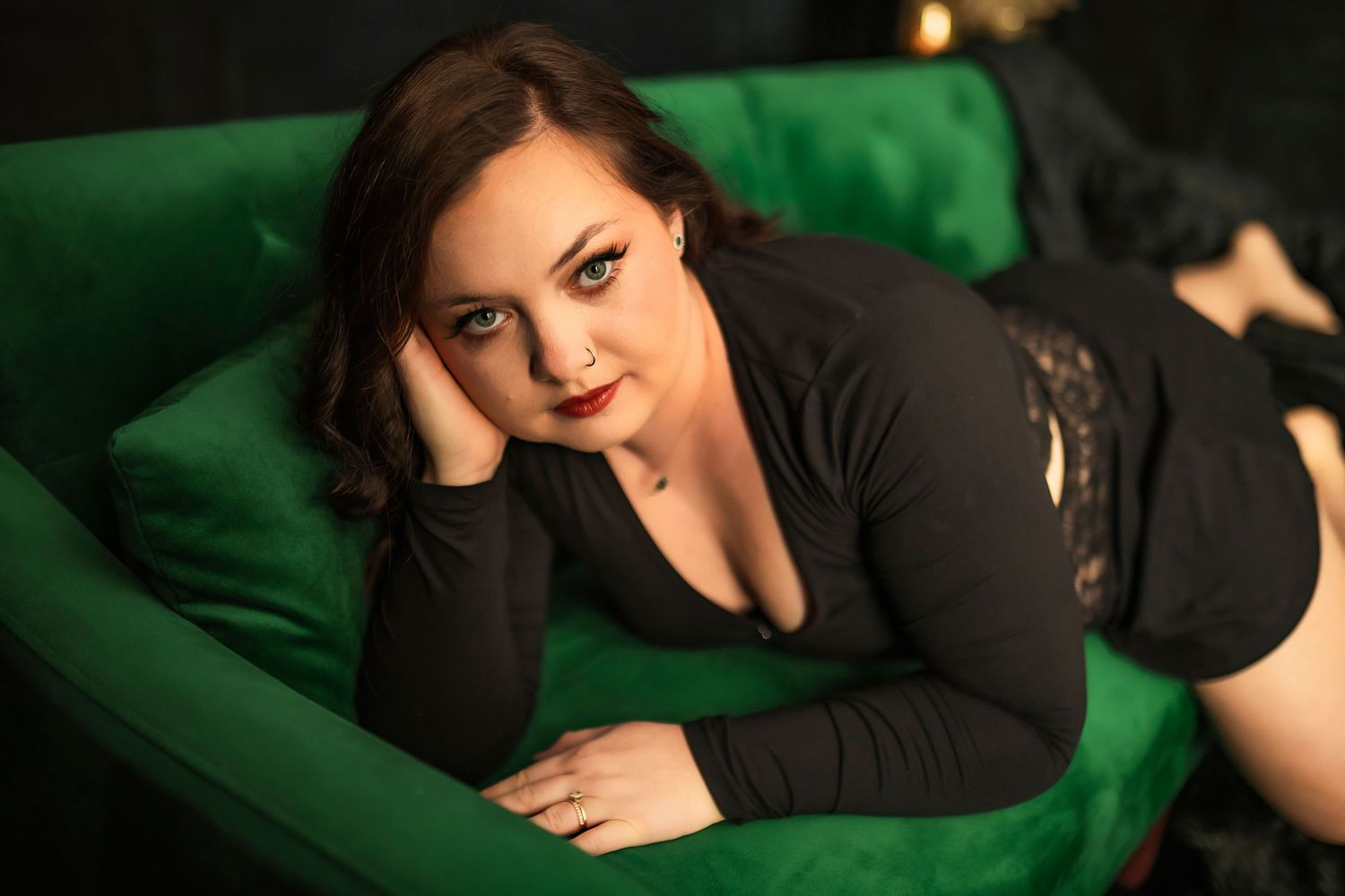 A person wearing black lounges seductively on an emerald green velvet couch in a dark room.