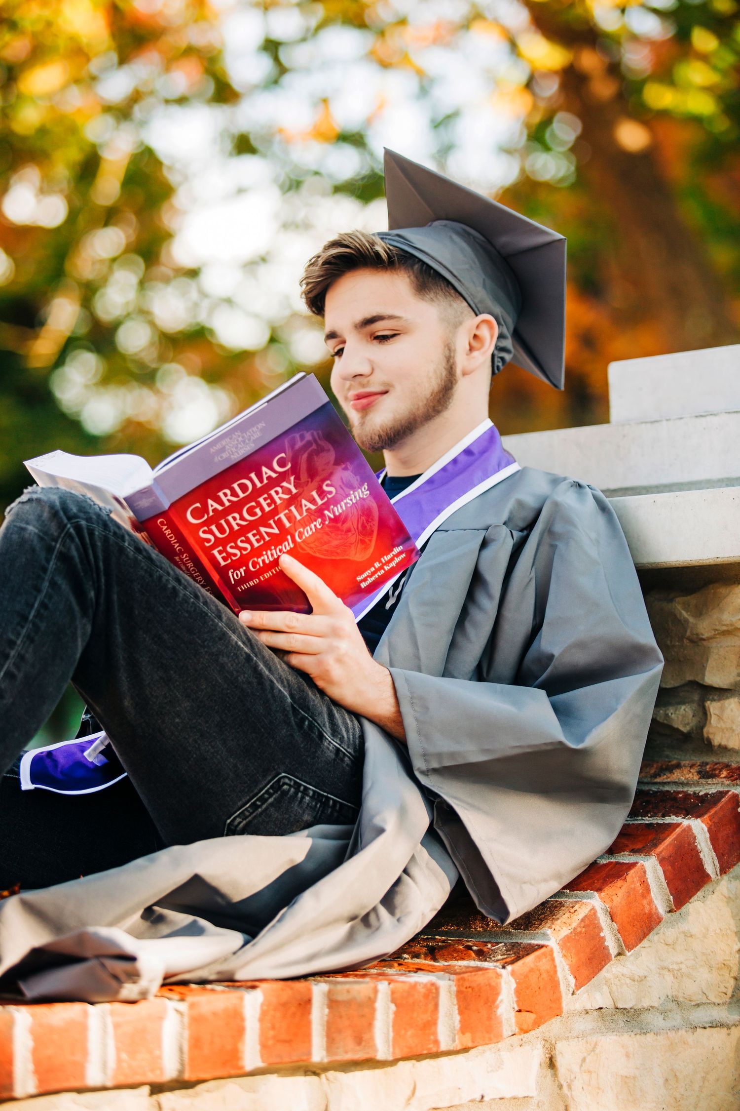 Graduate in cap and gown reading textbook while relaxing on brick wall outdoors.