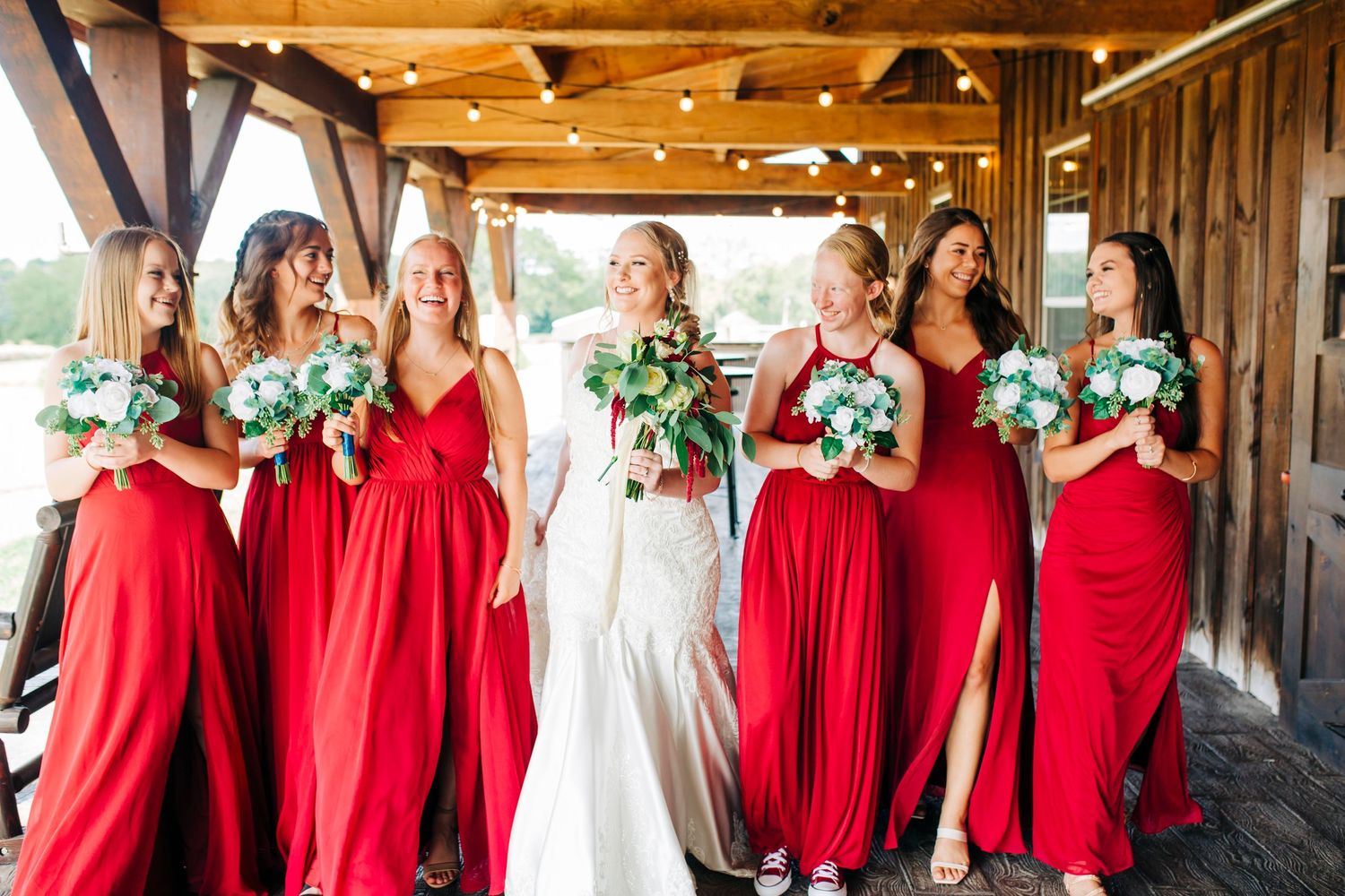 Bridesmaids in flowing red dresses stand together for a group photo in a rustic barn setting.