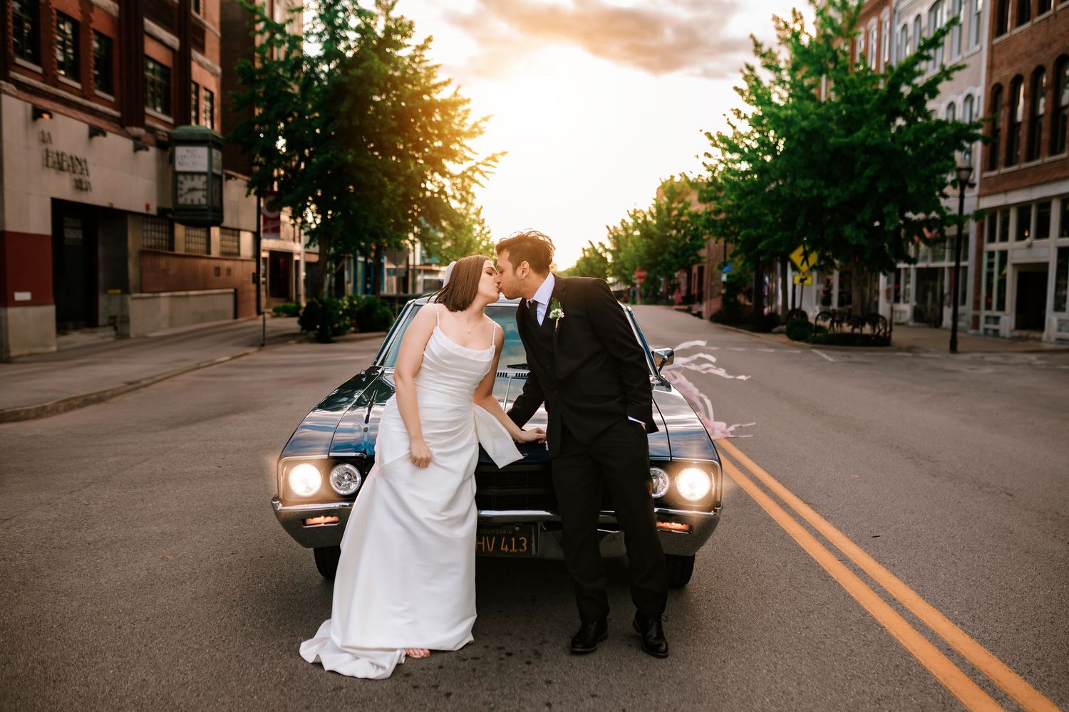 A happy couple leaning against a classic car in formal attire on an urban commercial street in downtown Springfield Missouri at sunset.