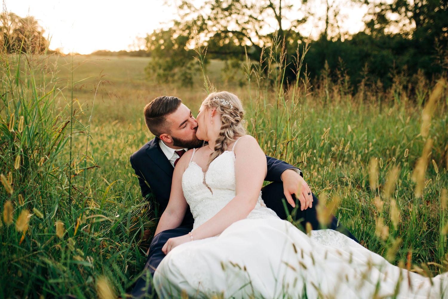 Newlyweds share intimate moment while sitting in tall grass at sunset.