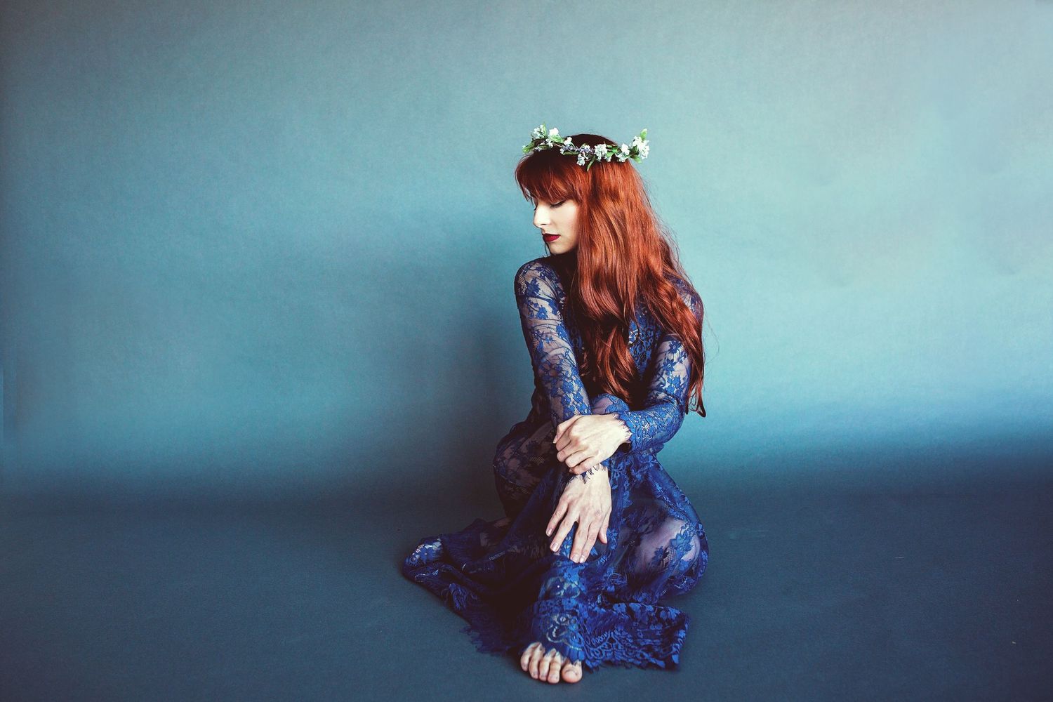 Person in blue lace dress sits on floor wearing floral crown against turquoise backdrop.