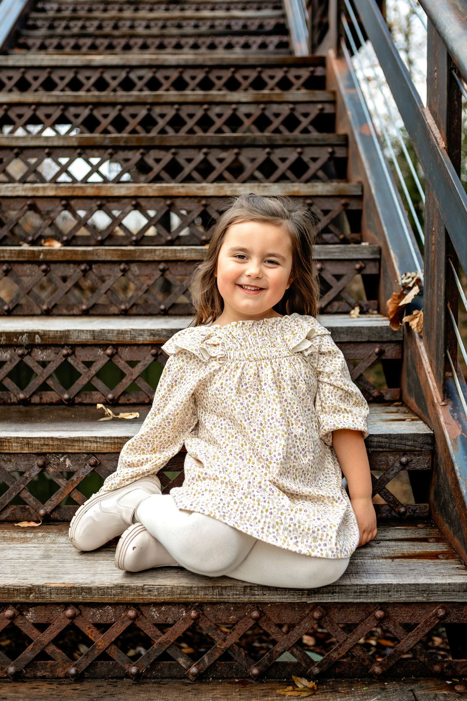 A seated child in a floral patterned dress and white leggings poses on decorative metal stairs outdoors.