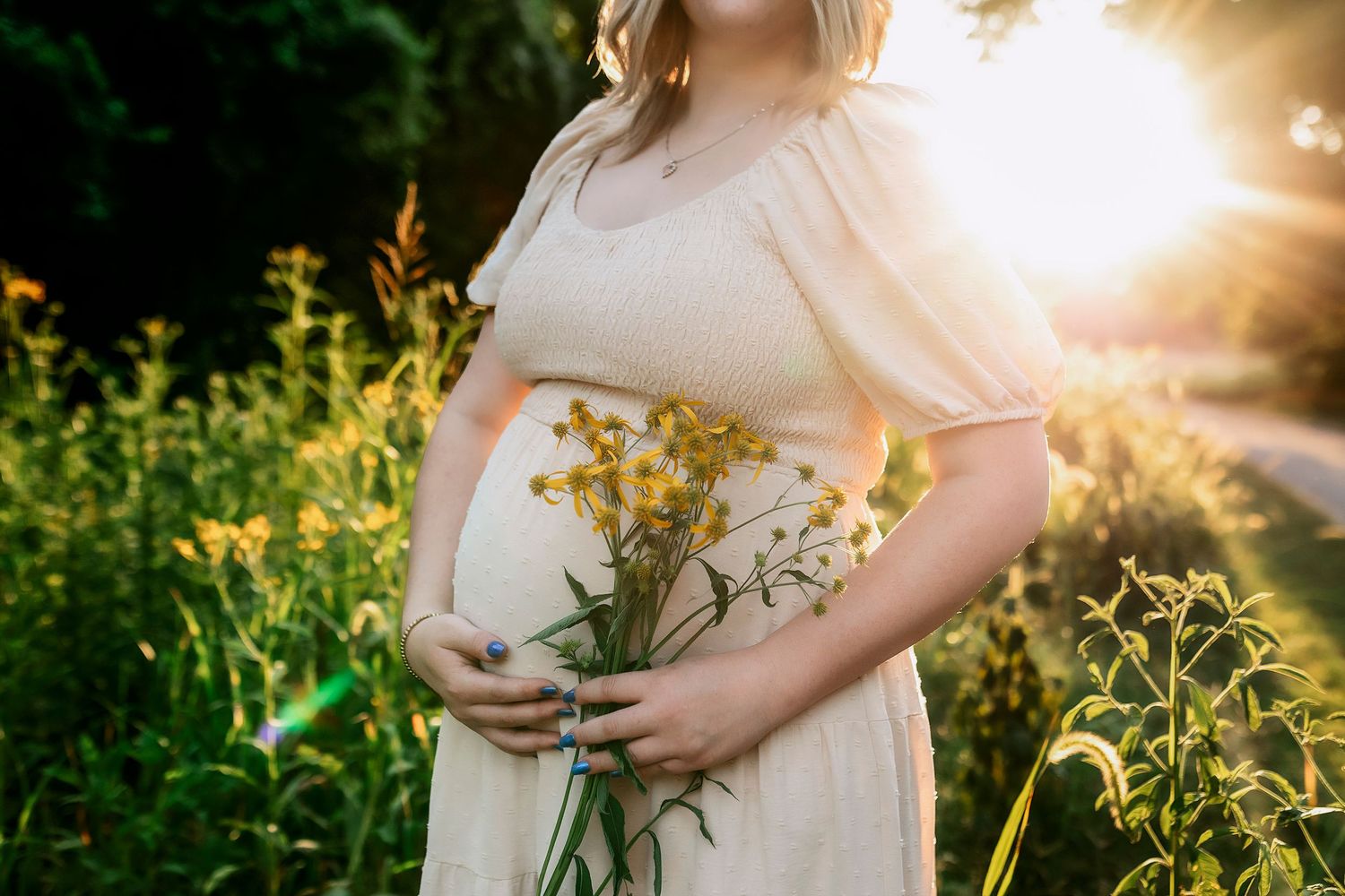 A pregnant woman in white dress holds yellow wildflowers in a sunlit meadow at golden hour.