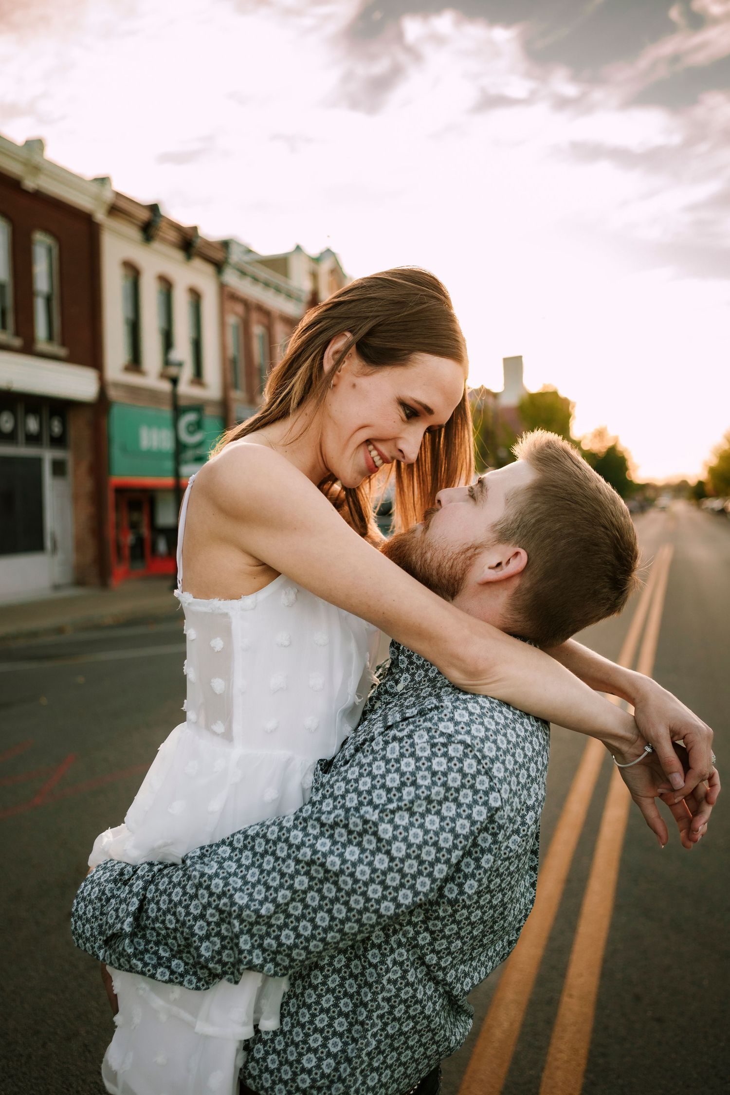 A romantic couple embraces and laughs together on an empty street at sunset, with the woman wearing a white dress.