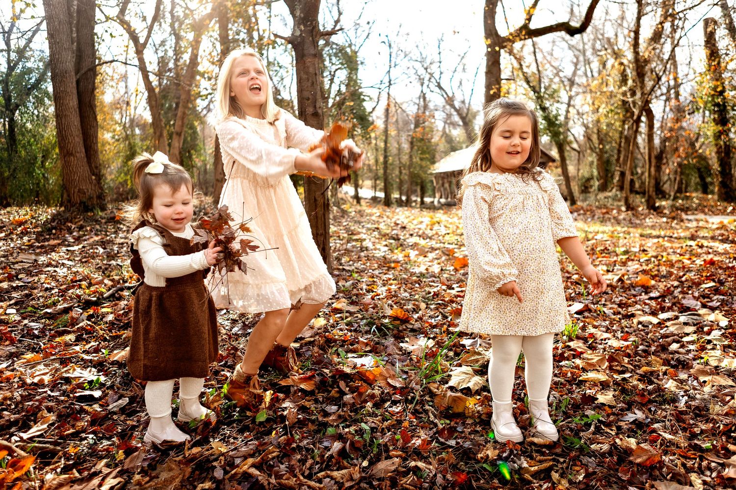 Three children play in fallen autumn leaves wearing vintage style dresses in a wooded outdoor setting.