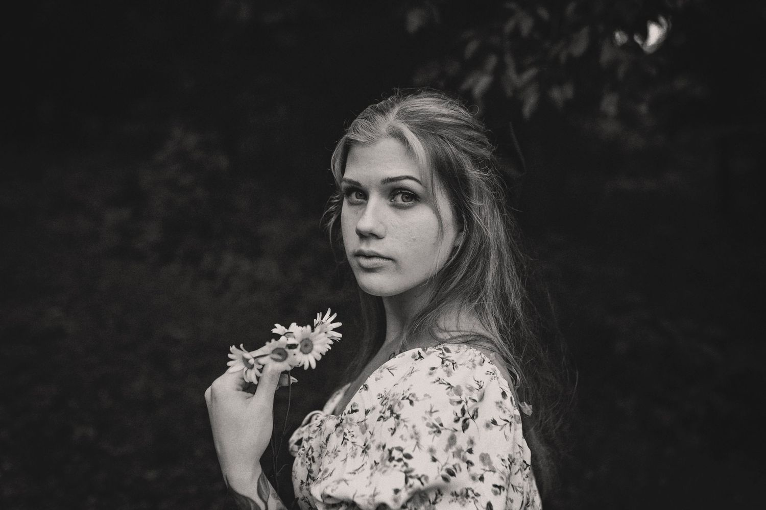 Black and white portrait of a person in floral dress holding flowers while looking away against dark foliage background.