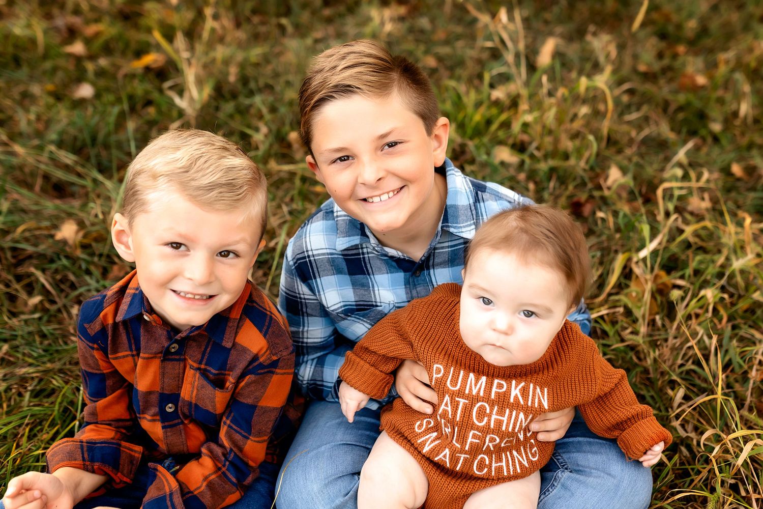 Three young children wearing plaid and autumn colors pose together outdoors in fall foliage.