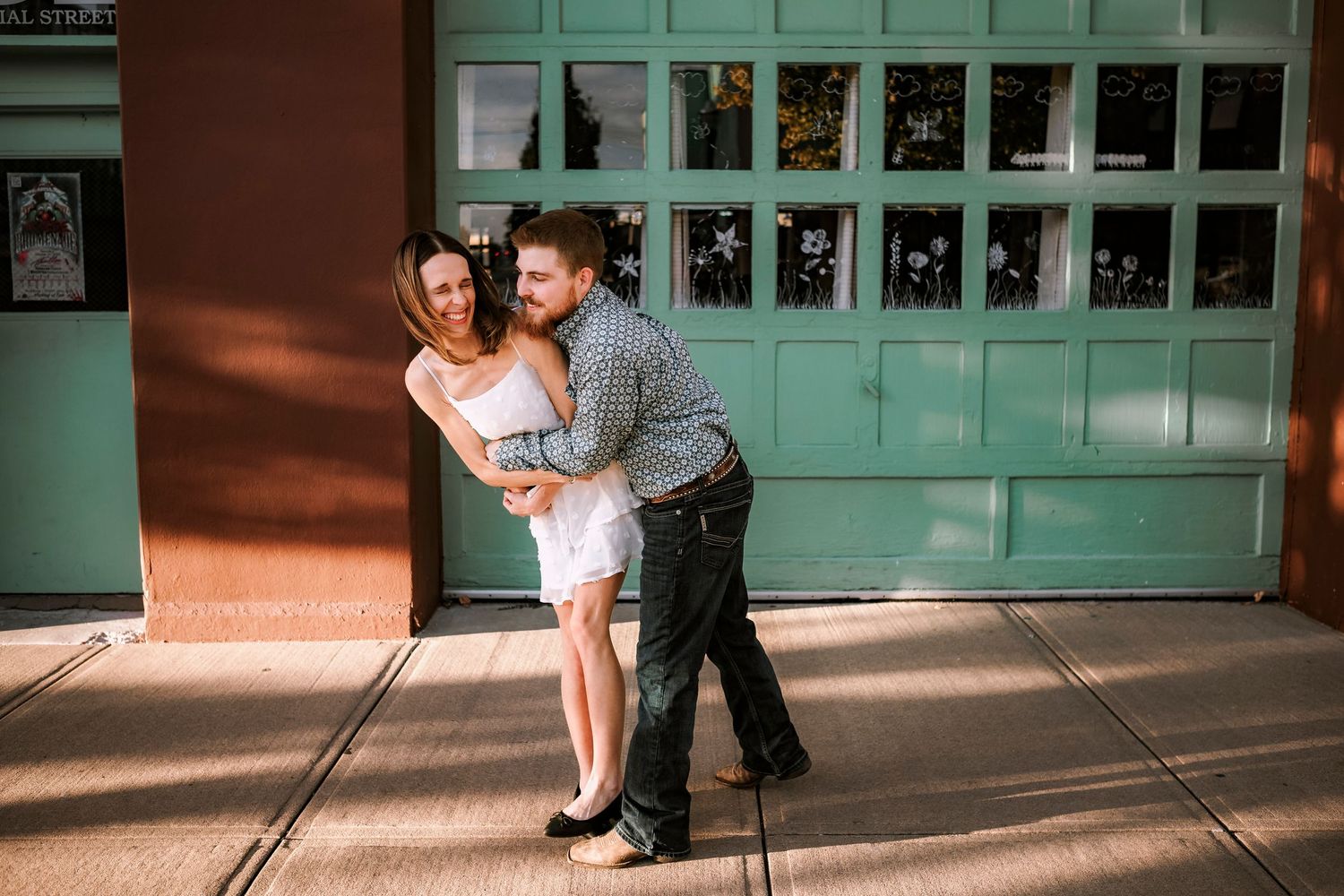 A couple embraces in front of a vintage turquoise garage door while dancing on a sunlit sidewalk.